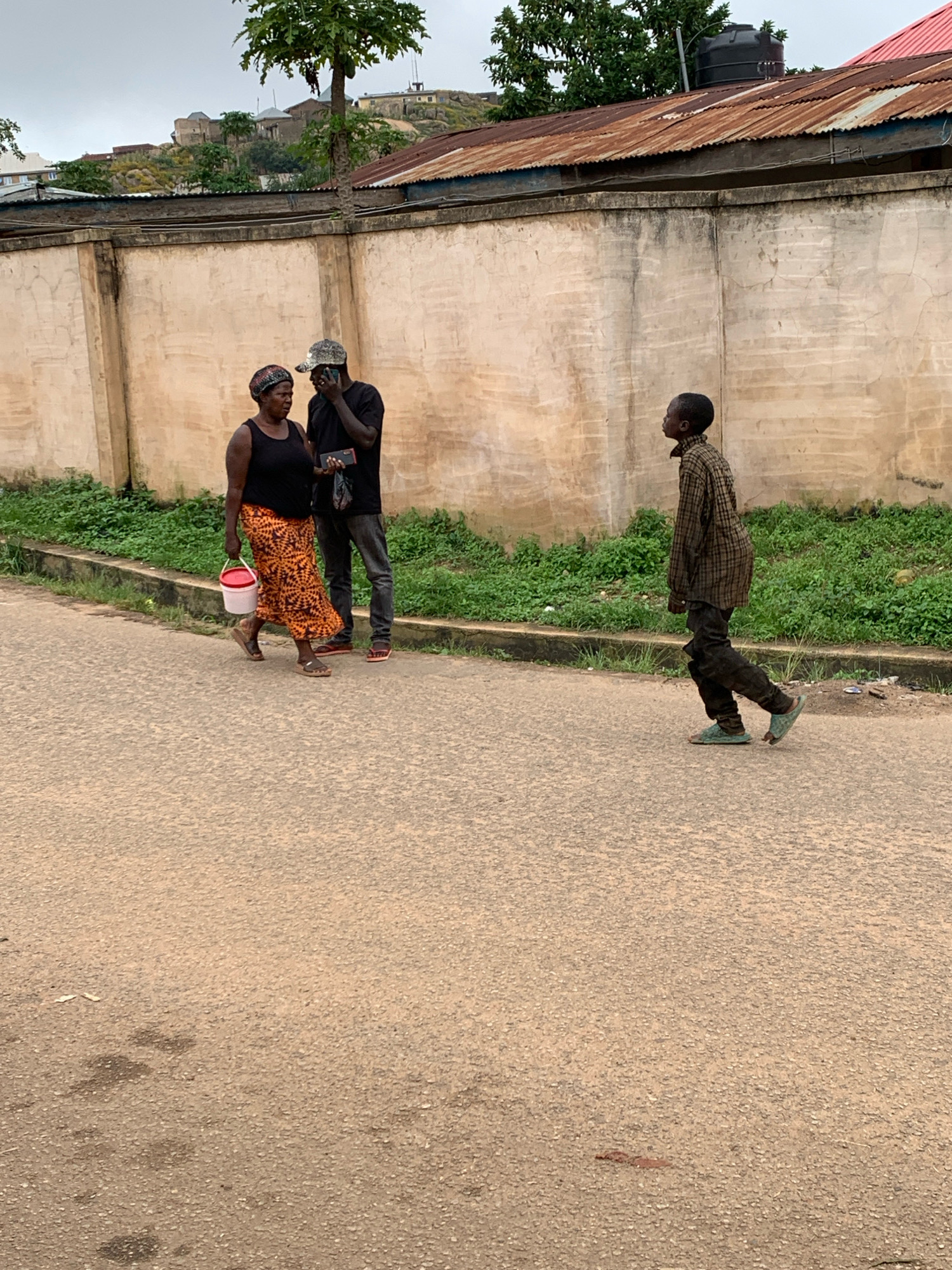 In Jos, Nigeria, on a paved road in front of a textured, pale wall, three individuals are visible. To the left, a woman wearing a black tank top, a patterned orange and brown skirt, and a headscarf, walks while carrying a red and white bucket in her left hand. Beside her, a man in a black t-shirt and dark pants, wearing a camouflage baseball cap, is looking at his phone. To the right, a young boy in a plaid shirt and dark pants walks away from the camera, wearing sandals. Green vegetation grows along the base of the wall. In the background, a weathered, corrugated metal roof is visible, and beyond that, buildings and structures are situated on a rocky incline under an overcast sky.