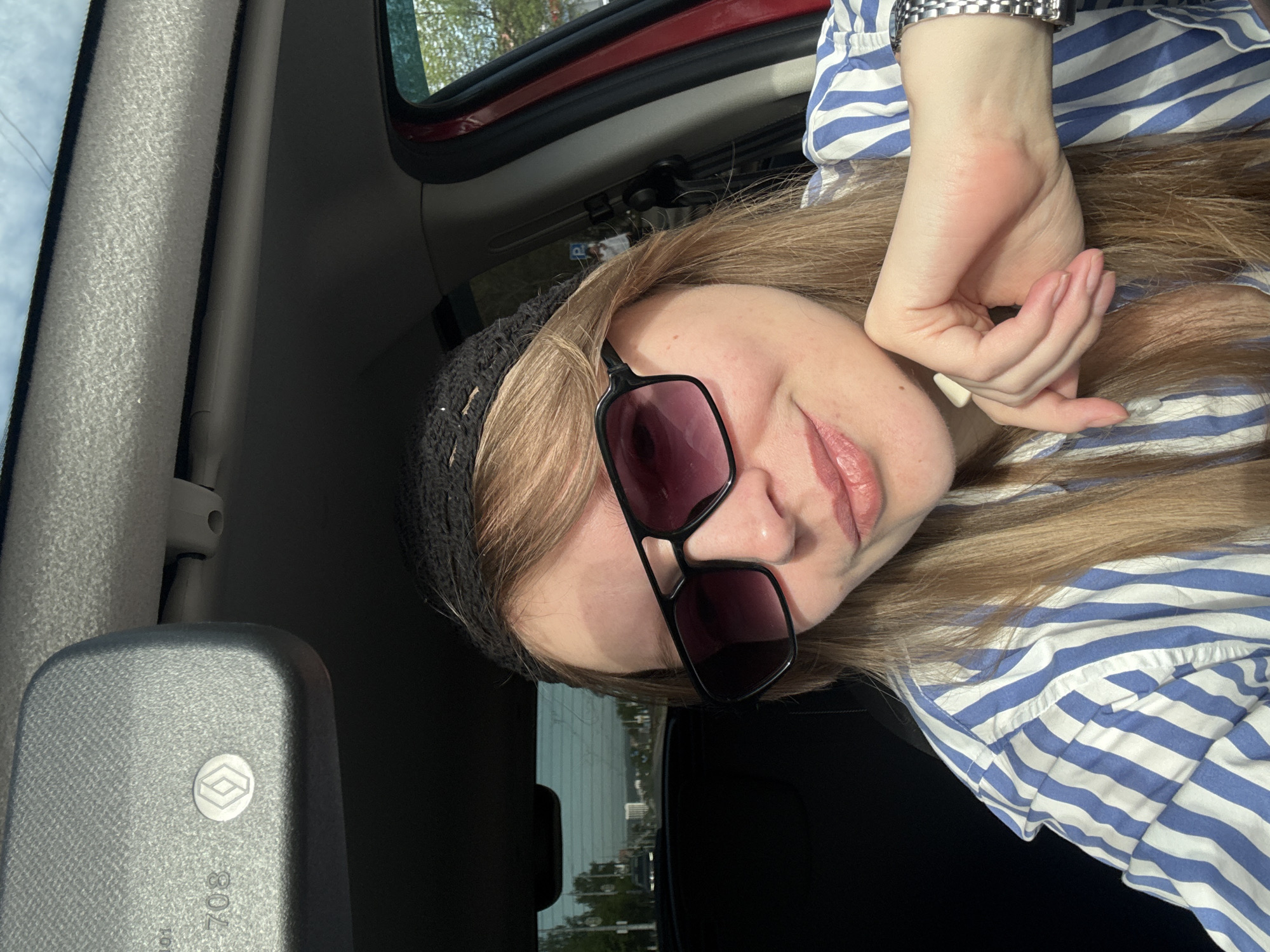 The image shows a woman in a vehicle, likely a car. She is wearing a blue and white striped shirt, a dark beanie, and sunglasses. Her face is partially visible, and her hand is near her face. The vehicle appears to be moving, as a cityscape is visible through the window. A car window and interior elements are visible. Based on the presence of the "708" marking on the car's interior, the location is Izhevsk, Russia.