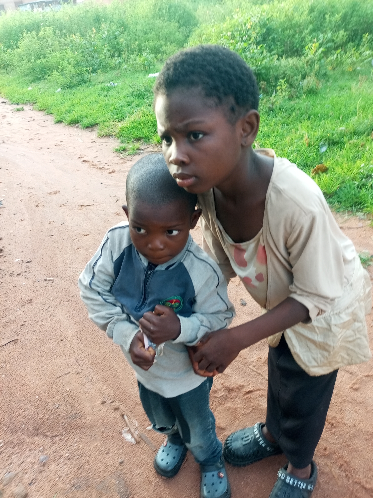 Two young children are standing on a dirt path with green grass and vegetation in the background. The child on the left is wearing a grey and blue jacket with jeans and blue Croc-style shoes. This child is holding a small object, possibly a piece of candy or paper, in their right hand. The child on the right is wearing a light-colored shirt and black pants, also with black Croc-style shoes. This child has their arm around the other child's shoulders. The scene appears to be outdoors, possibly in a rural or suburban area of Tempure, Nigeria. The lighting suggests it is daytime.