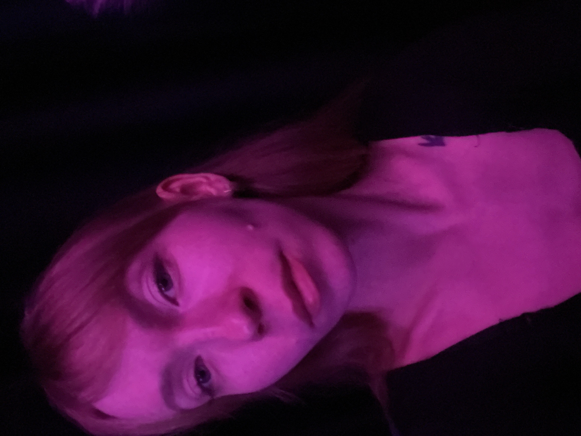 A close-up photograph shows a fair-skinned person, illuminated with pink light, against a dark background. The person is tilted to the side, with their face and upper chest visible. The eyes are open, and the lips are closed. There appears to be short red hair, and there is a tattoo on the upper chest. The lighting creates a predominantly magenta hue across the subject. No specific location or setting details relating to the city of Perm, Russia, are evident.