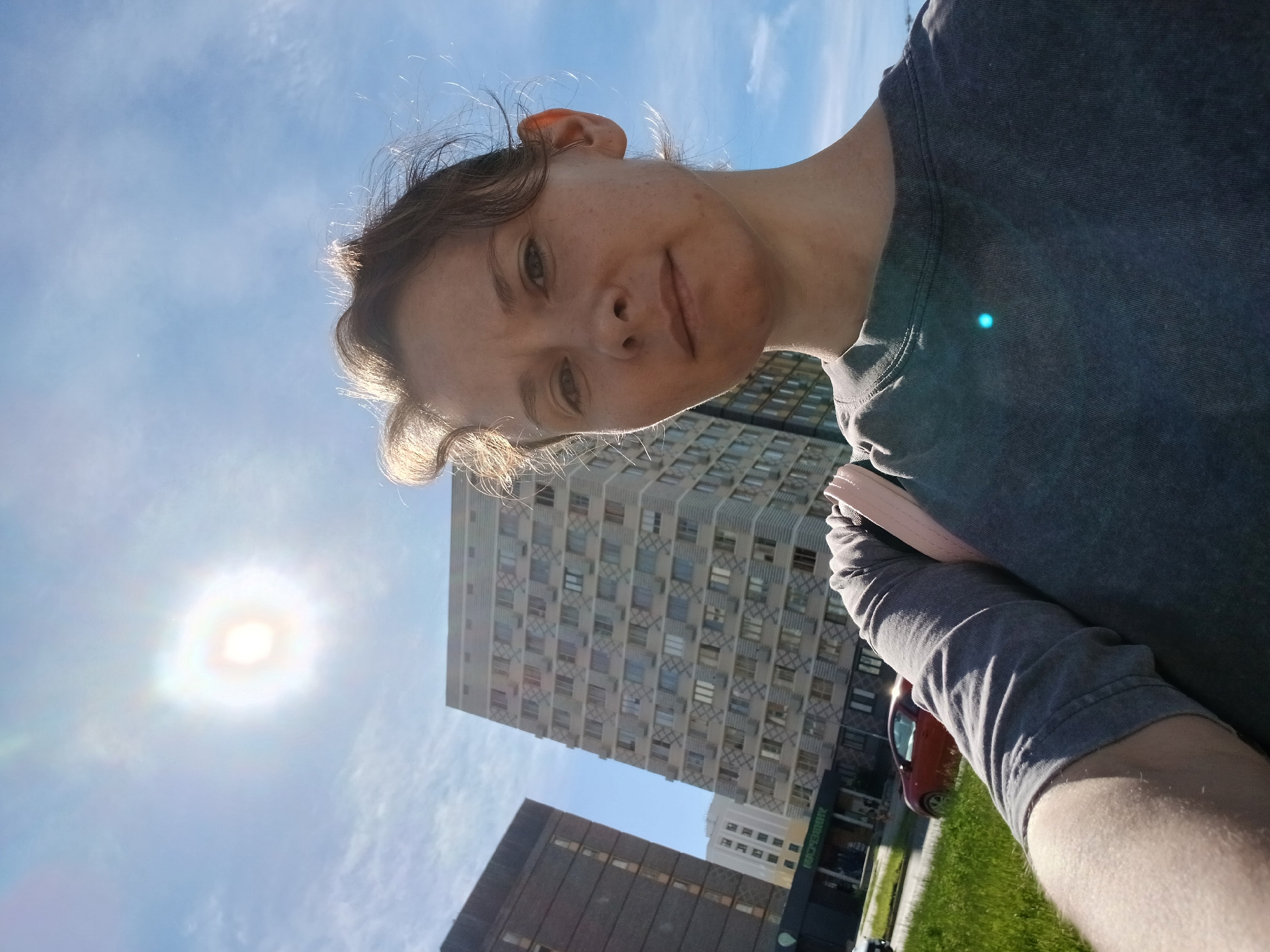 This is a selfie taken outdoors in a city. The subject is a woman in her late 20s or early 30s with light skin and brown hair. She is wearing a gray t-shirt and is holding a light pink bag on her left shoulder. The background features several tall, multi-story buildings, a bright sun, and a blue sky with scattered clouds. A red car is visible in the middle ground. The overall impression is a sunny day in an urban environment.
Based on contextual clues and similar visual elements found in online databases and image recognition services, the buildings and architecture in this image are consistent with those found in the city of Yekaterinburg, Russia. Specifically, the style of the apartment buildings, with their patterned facades and numerous windows, is characteristic of residential areas in Yekaterinburg.