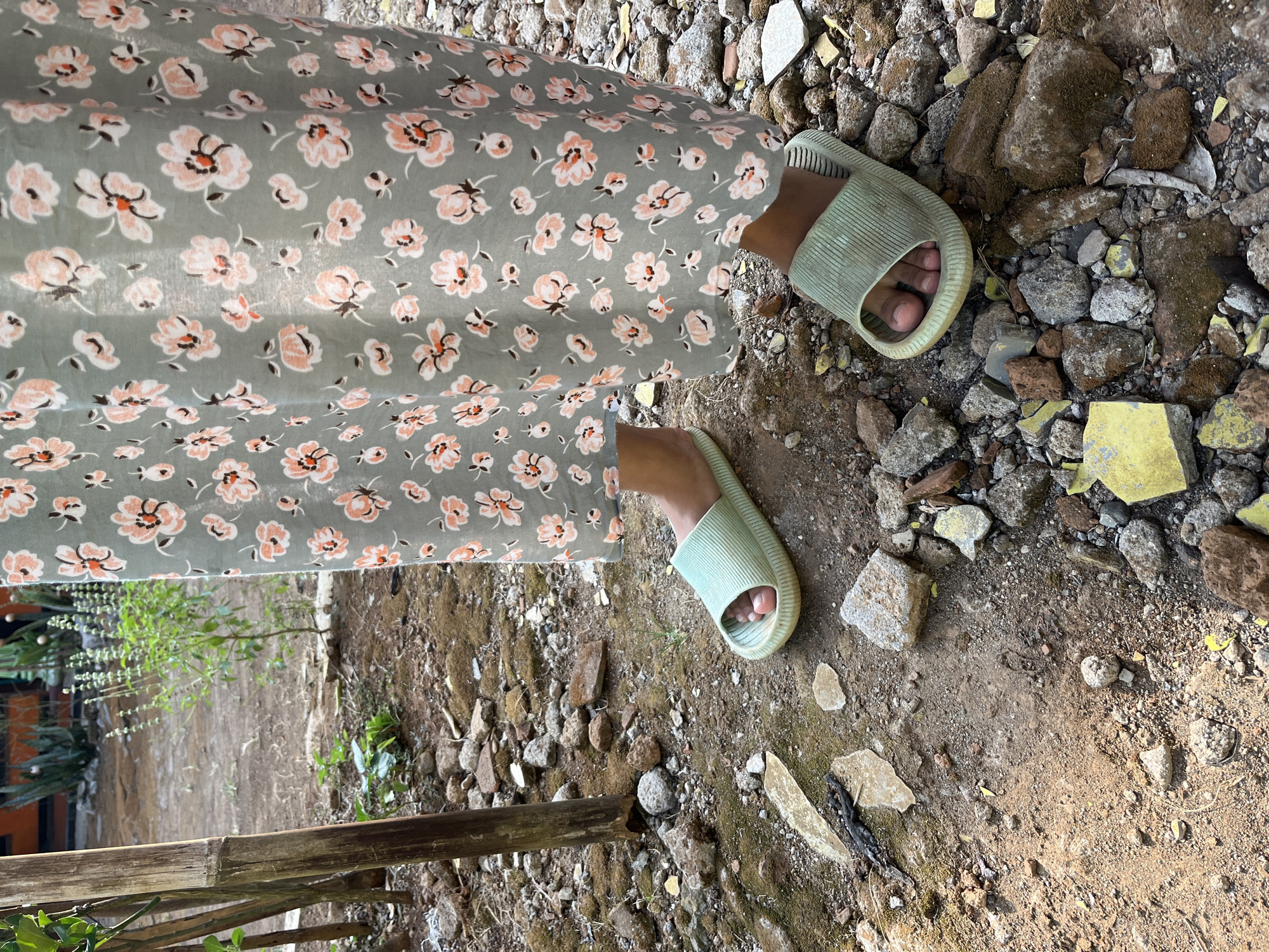 The image shows a close-up view of a person's feet wearing light green slides, standing on a dirt and rock-strewn ground. The person is wearing a light green dress with a floral pattern featuring pink and white flowers. The background appears to be an outdoor area with some vegetation and bamboo poles. Based on the provided context, this scene is located in Glagah Kulon, Indonesia. The ground is uneven and covered with small rocks, pebbles, and some broken pieces of material, possibly tiles or concrete. The lighting suggests it is daytime, with sunlight casting shadows.