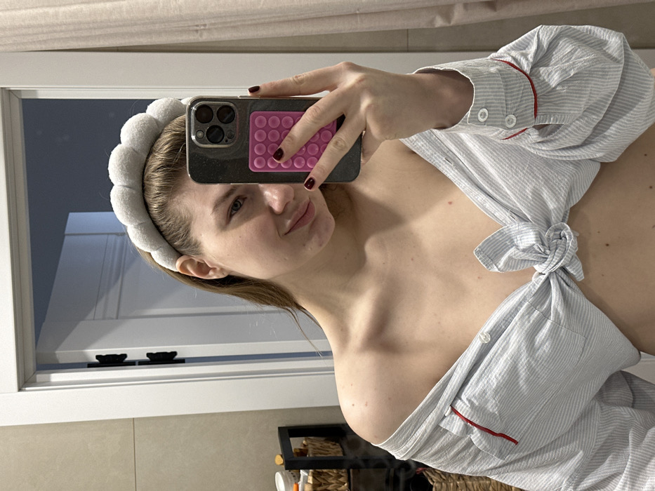 A young adult female is depicted in a mirror selfie, located in what is described as the city of Grebeni, Russia, within an indoor setting resembling a bathroom or dressing area. She has fair skin, light brown hair pulled back by a grey, cloud-textured headband, and a slight smile. She wears a light blue and white striped, long-sleeved button-up shirt, unbuttoned and tied at the midriff, exposing her abdomen and shoulders. The shirt cuffs feature red trim. Her left hand holds a dark-colored smartphone, equipped with a bright pink, rectangular textured grip on its back, positioned to capture her reflection. Her fingernails are painted dark red. In the background reflection, a light-colored door with dark hinges is visible to her right. To her left, a dark-framed shelving unit holds various items, including a wicker basket and several containers. The walls are a light, neutral color.