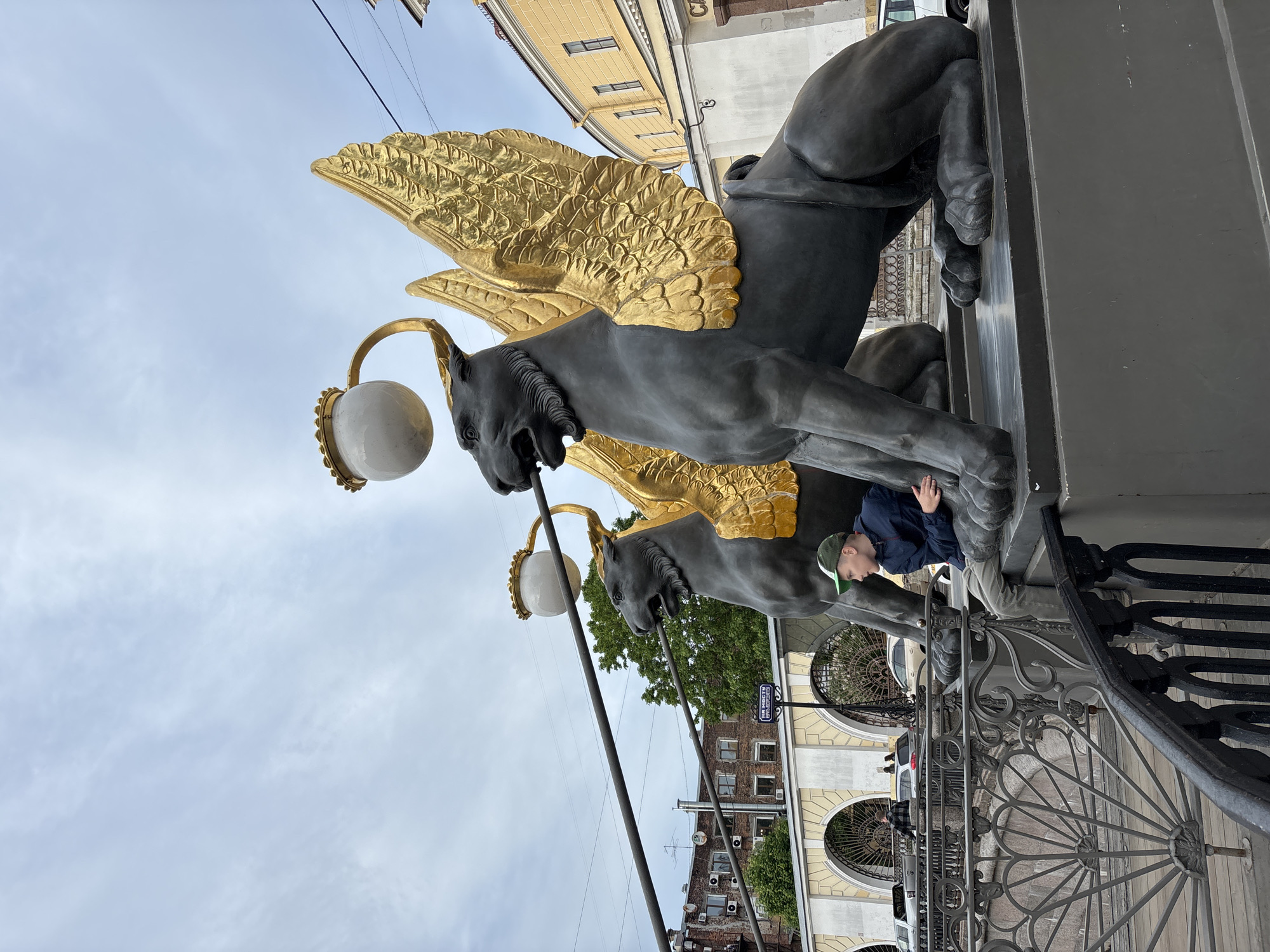 The image, taken in Saint Petersburg, Russia, features two dark grey statues of winged gryphons with prominent golden wings. Each gryphon holds a white spherical lantern, part of an ornate street lamp design. The photo is rotated to be portrait orientation, showing the gryphons from a low-angle perspective, emphasizing their grandeur against an overcast, light blue sky.
A young person, possibly a child or teenager, wearing a blue jacket and a green baseball cap, is visible in the lower right quadrant, leaning against the base of one of the gryphon statues. They appear to be observing the statue or posing alongside it.
In the background, several buildings with yellow, cream, and darker brick facades are visible, along with windows and some air conditioning units. Green foliage from trees can also be seen. An ornate black metal railing, likely part of a bridge, spans the bottom right of the frame. A blue sign with white text, "НА МОСТУ" (meaning "ON THE BRIDGE" in Russian), is visible in the middle-ground, further indicating the bridge setting. The overall weather appears cloudy, suggesting an overcast day.