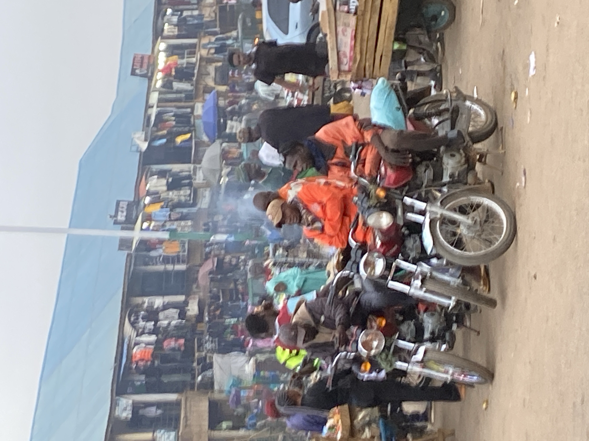 This image captures a vibrant and bustling outdoor market scene in Bukuru, Nigeria. The setting is characterized by numerous market stalls or small shops lining what appears to be a dusty, unpaved street or open area. The sky above is hazy or overcast.
In the foreground and mid-ground, a prominent feature is a gathering of several motorcycles, which are a common mode of transport in the region (often called "okadas"). Some motorcycles are parked, while others have riders on them. Many people are visible, congregating around these motorcycles and moving through the market. Several individuals are wearing bright orange attire, possibly indicating a uniform or a specific group, such as motorcycle taxi operators or market vendors. One person wears a light blue head covering. A visible plume of smoke or dust rises from the mid-ground, contributing to the busy atmosphere.
The background is densely packed with market structures, displaying various goods. Items resembling clothing are visible hanging in some stalls. Faintly visible text on signs, though inverted, includes words like "LADIES" and "ZARA," suggesting the types of products sold. Amidst the stalls, a blue umbrella and a lighter-colored umbrella are open, likely providing shade or protection from light elements. A partially visible white car or van, along with another smaller vehicle or cart, also adds to the market's activity. The scene conveys a sense of energetic daily commerce and transportation in a community hub.