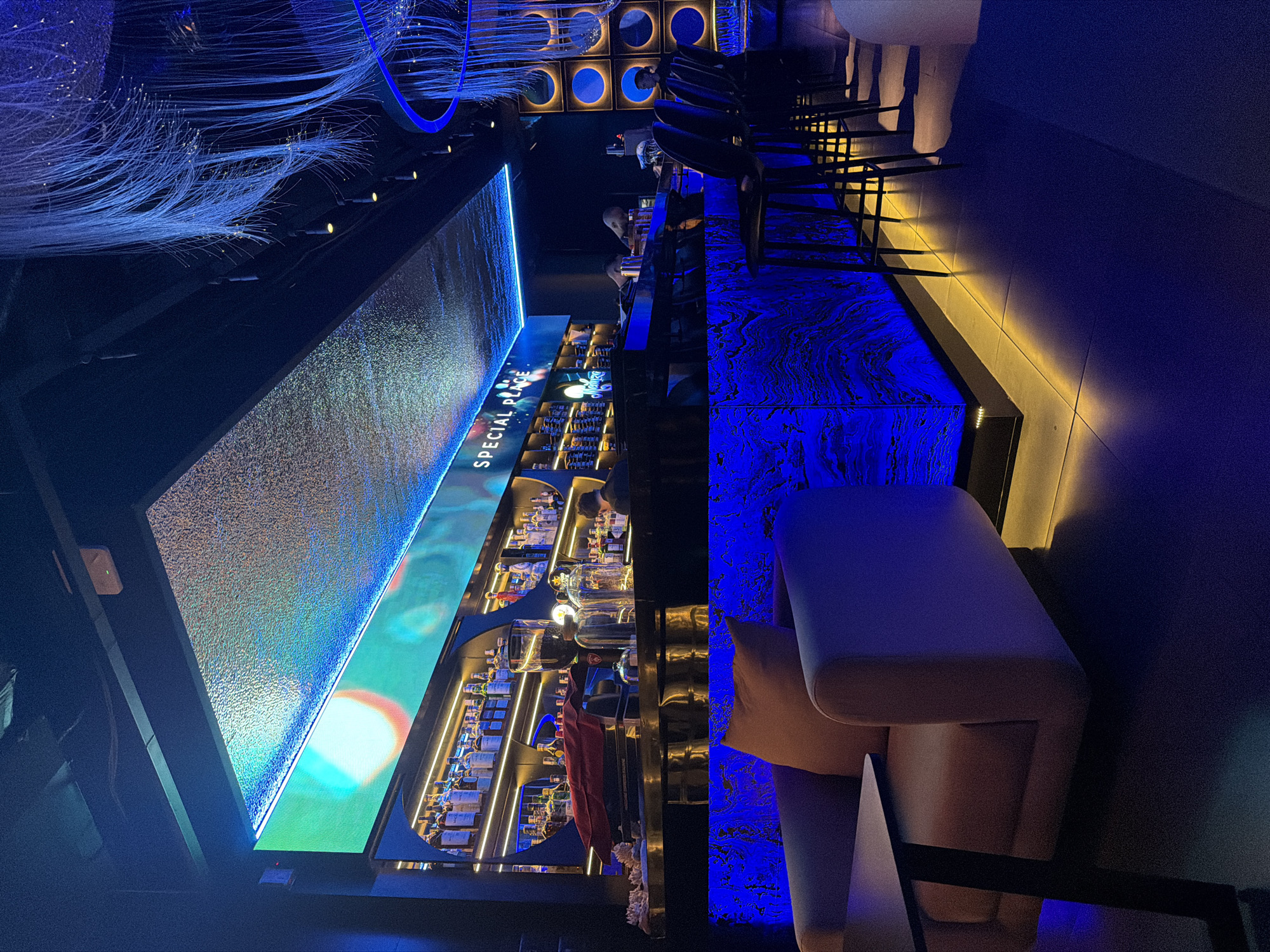 The image depicts the interior of a modern, dimly lit bar, possibly a nightclub, with vibrant blue and yellow lighting. In the foreground, there are two plush chairs with a small table to the left, bathed in a soft orange glow from the chair cushions and blue from the surrounding lights. To the right, a sleek bar area is visible, featuring a marble-patterned counter illuminated by bright blue LED lights. Several bar stools with black seats and thin metal legs are neatly arranged along the counter. Behind the counter, shelves are stocked with various bottles of alcohol, illuminated by warm yellow lights. A large LED screen displaying abstract colorful graphics is mounted above the bar, with the words "SPECIAL PLACE" illuminated in white. In the background, intricate fiber optic light installations hang from the ceiling, casting a delicate glow. The environment suggests a sophisticated and atmospheric venue. The location information provided states this scene is in Nha Trang Ward, Vietnam.