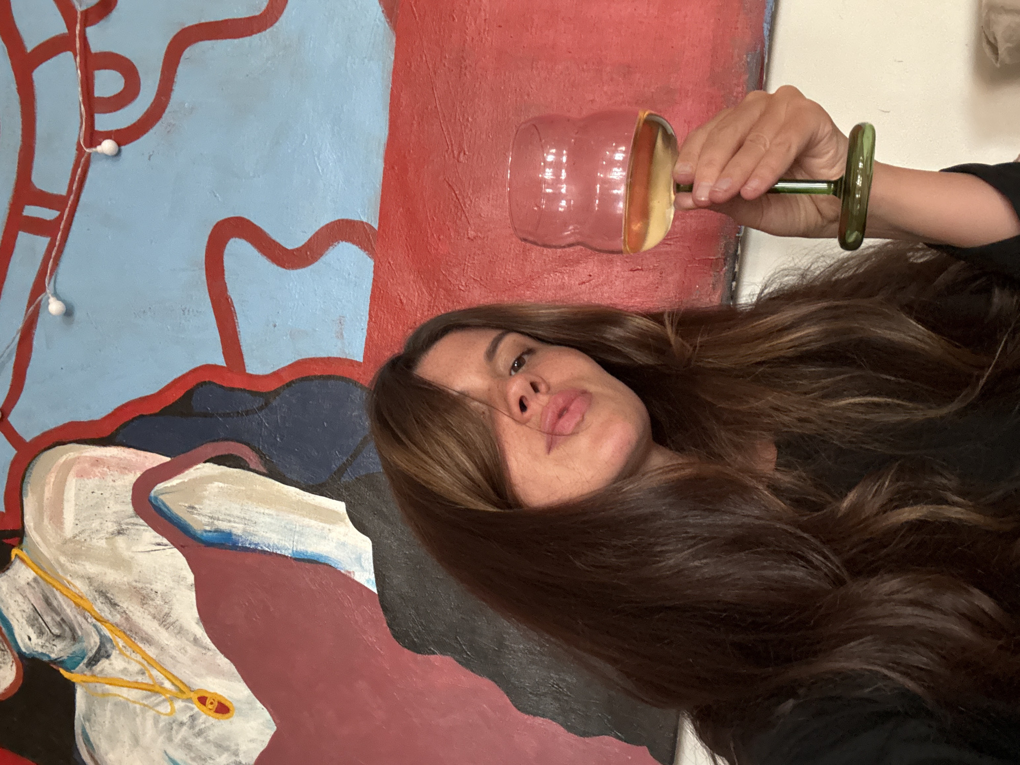 An angled photograph shows a woman holding a unique drink glass, set against a large, colorful abstract painting in what appears to be an indoor space. The woman, whose head and upper torso are visible, has long, wavy dark brown hair that frames her face and flows over her shoulders. She looks towards the camera with a slightly pouty or contemplative expression, her lips parted.
She holds an artistic glass with a ribbed, pink-tinted bowl, a slender green stem, and a metallic gold-colored base. A light-colored liquid is visible inside the glass.
The background is dominated by a vibrant abstract painting featuring broad sections of light blue, deep red, dark blue, white, and maroon. Organic, flowing red lines are painted over the light blue area. Below, abstract shapes in white, maroon, and dark blue are visible, with a thin yellow line and a small red detail on one of the white forms. Small, spherical white lights or beads hang vertically on the upper left section of the painting. The scene suggests an artistic or personal setting, located in Krasnoyarsk, Russia. There is no visible text.