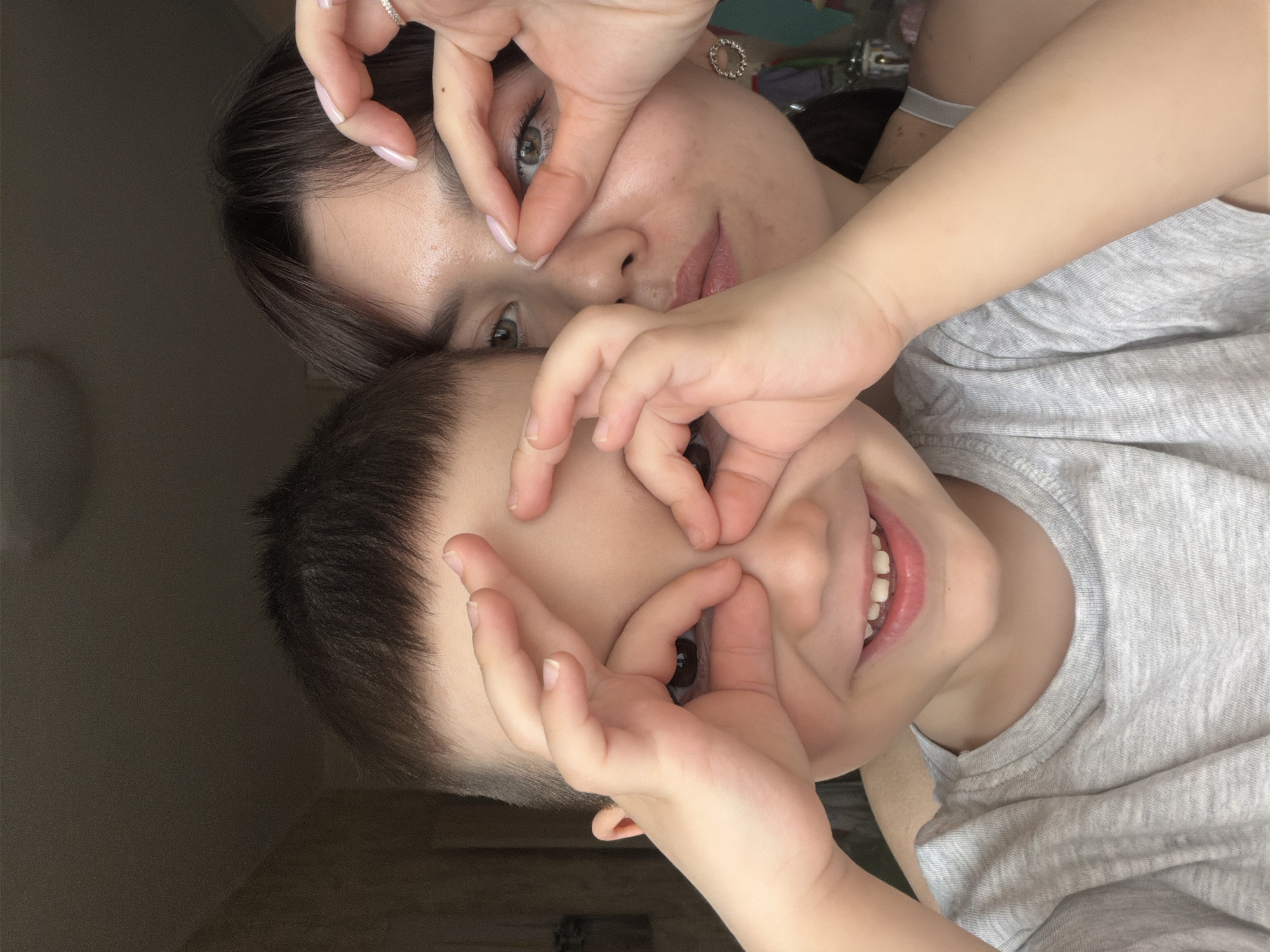 A selfie shows a woman and a young boy making circles around their eyes with their hands. The woman is at the top of the frame, and the boy is below her. Both have fair skin. The woman has dark brown hair, green eyes, and pink lipstick, with a ring on her left ring finger. The boy has short brown hair and is smiling, revealing his teeth. Both are wearing gray t-shirts. The background is blurred. The location cannot be determined from the image.