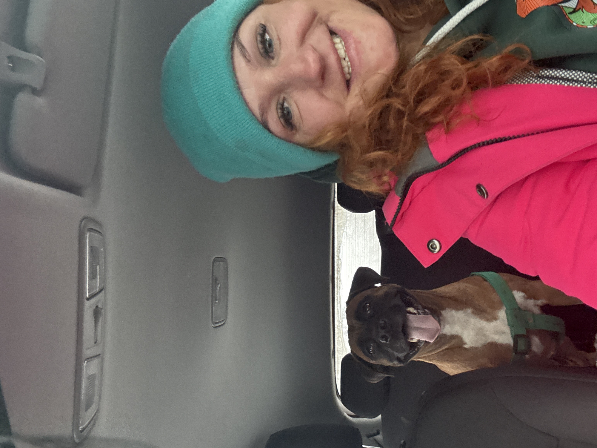 A selfie-style photograph, oriented vertically, shows a woman and a dog in what appears to be a car. The woman, at the top, wears a teal beanie and a pink jacket. She is smiling. Beneath her, a boxer dog with a green harness is visible. The interior of the car is visible, including the roof with interior lights and the passenger seat. The specific location in Chaikovsky, Russia is not discernible from the visual information provided.