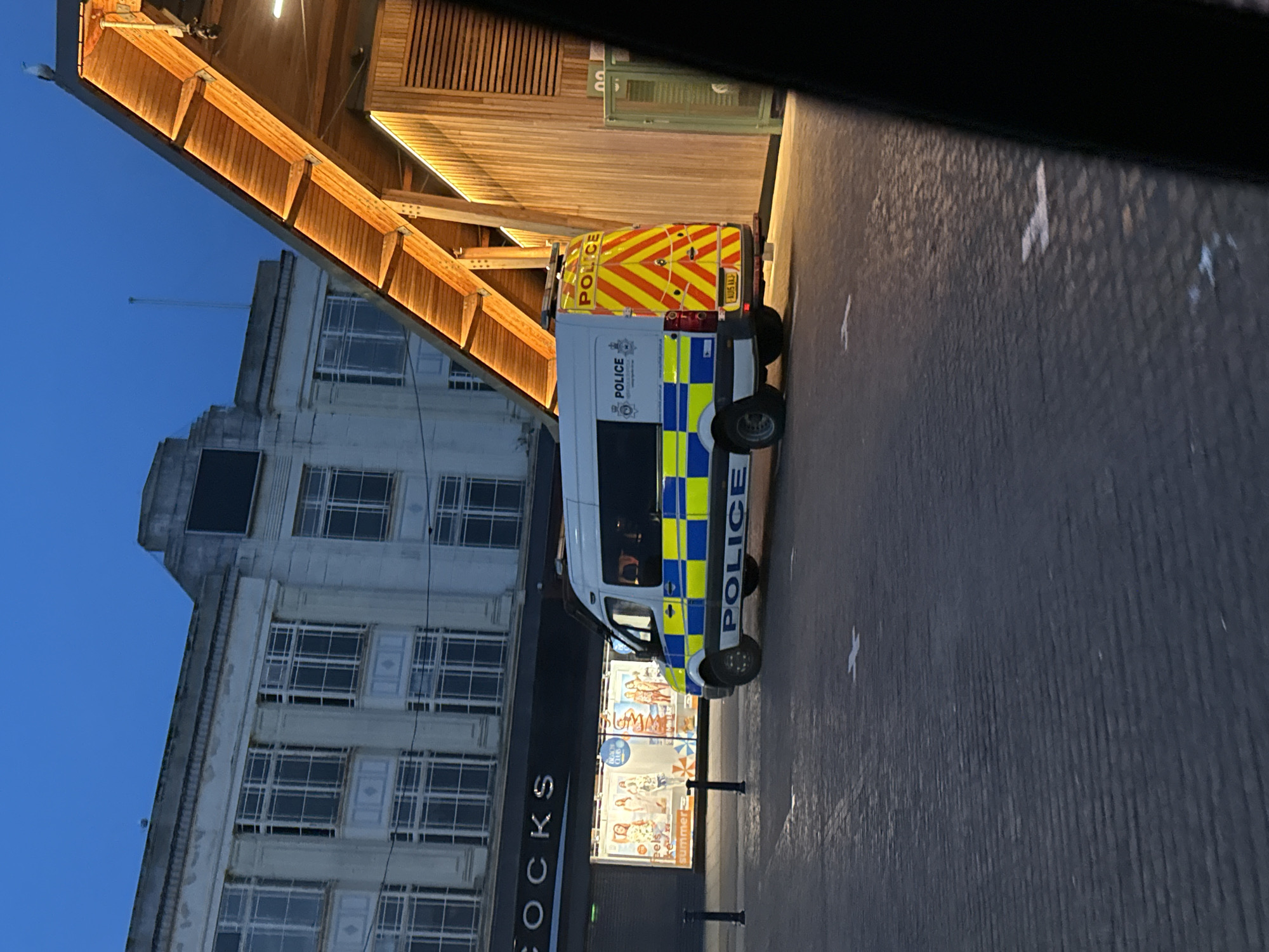 A police van is parked on a brick-paved surface in front of a multi-story building with "COCKS" signage. A wooden pedestrian bridge is overhead. The sky is blue. This is located in Great Yarmouth, United Kingdom.