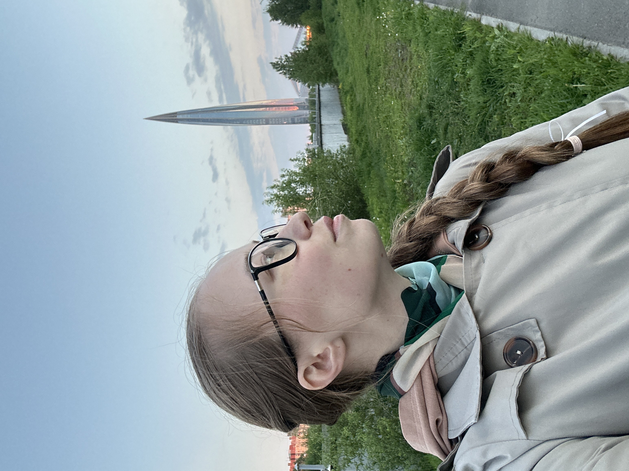 A young woman with fair skin, wearing black-rimmed glasses and a beige trench coat, is positioned in the foreground of the image. She has brown hair braided down her back, tied with a white and peach-colored elastic. The woman is looking upwards and slightly to the left.
In the background is the Lakhta Center, a skyscraper located in Saint Petersburg, Russia. It is recognizable by its modern, pointed design, and is lit by the setting sun. Green trees and grass occupy the middle ground, while the sky is a mix of blue and light orange hues, indicating either sunrise or sunset. The lighting suggests it is early morning or late evening. The focus is sharp, and the image is taken from a low angle, emphasizing the height of the Lakhta Center.