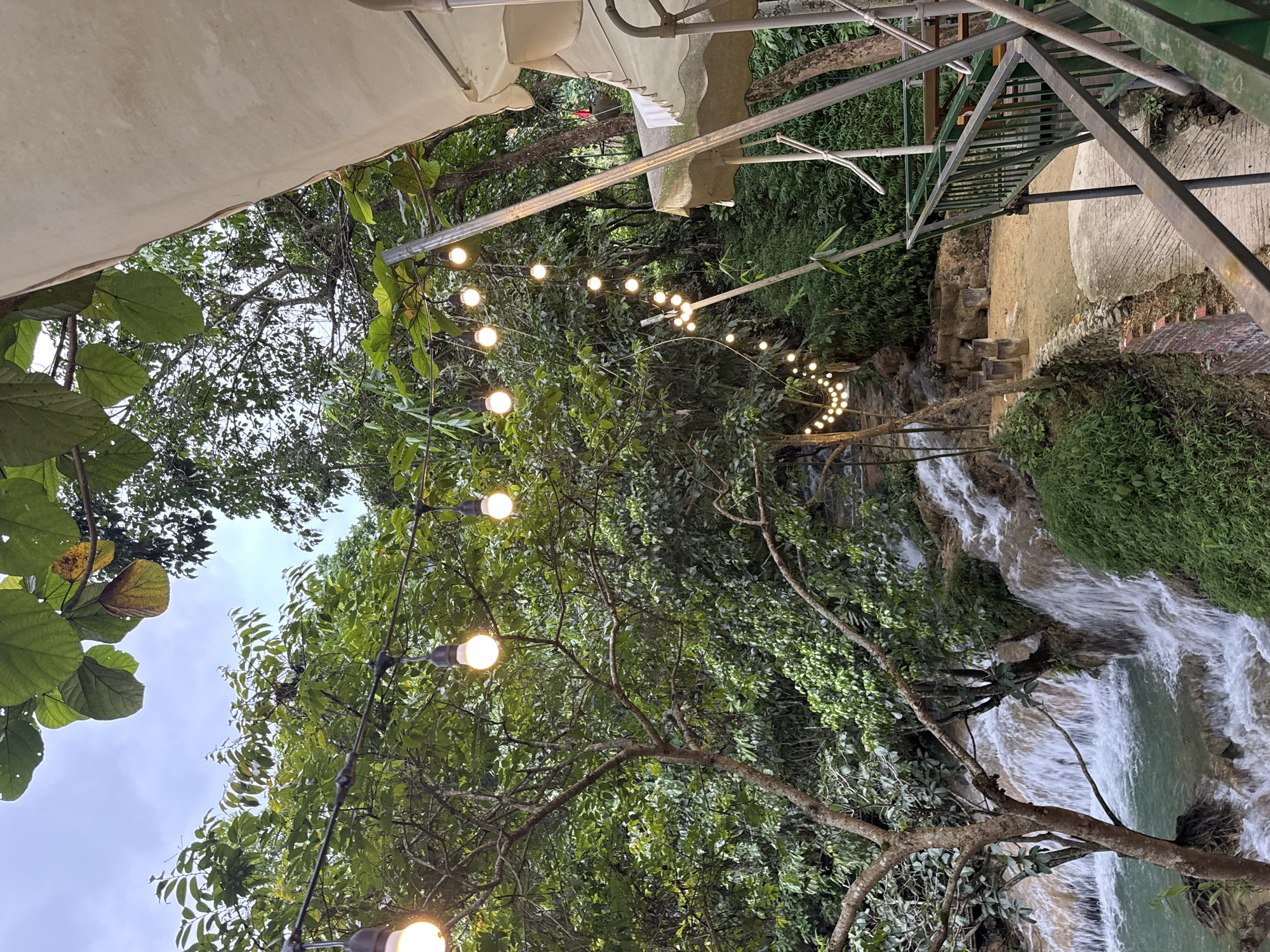 The image depicts an outdoor dining area adjacent to a natural waterfall. String lights illuminate a path leading towards the cascading water. A portion of a thatched roof and a metal railing are visible on the right side of the frame, suggesting a restaurant or cafe setting. The surrounding environment is lush with trees and greenery. The scene is located in Cổ Lũng Commune, Vietnam.