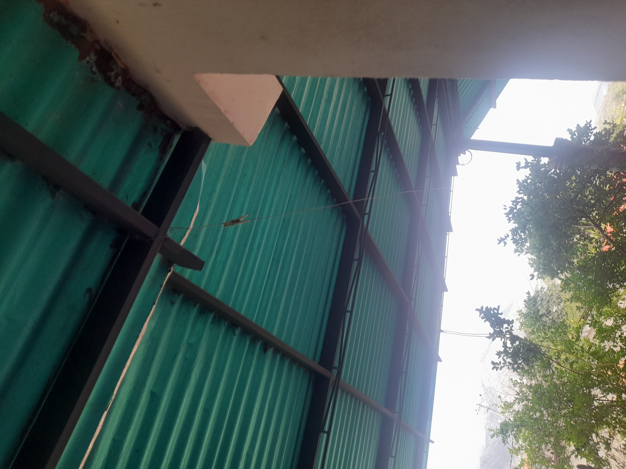 This image shows a view from under a green corrugated metal roof, supported by a dark metal frame. A beige concrete pillar is visible on the right side of the frame. The roof is partially covered in what appears to be dirt or debris along the edge where it meets the pillar. A thin string hangs from the roof structure, with a small, light-colored object resembling a clothes peg attached near the top. Through the gaps in the roof structure and to the left, lush green trees and vegetation are visible, with a hazy landscape of hills or mountains in the background under a bright sky. The image appears to have been taken outdoors. There are no people visible in the image. The location is identified as Santa Ana, Venezuela.