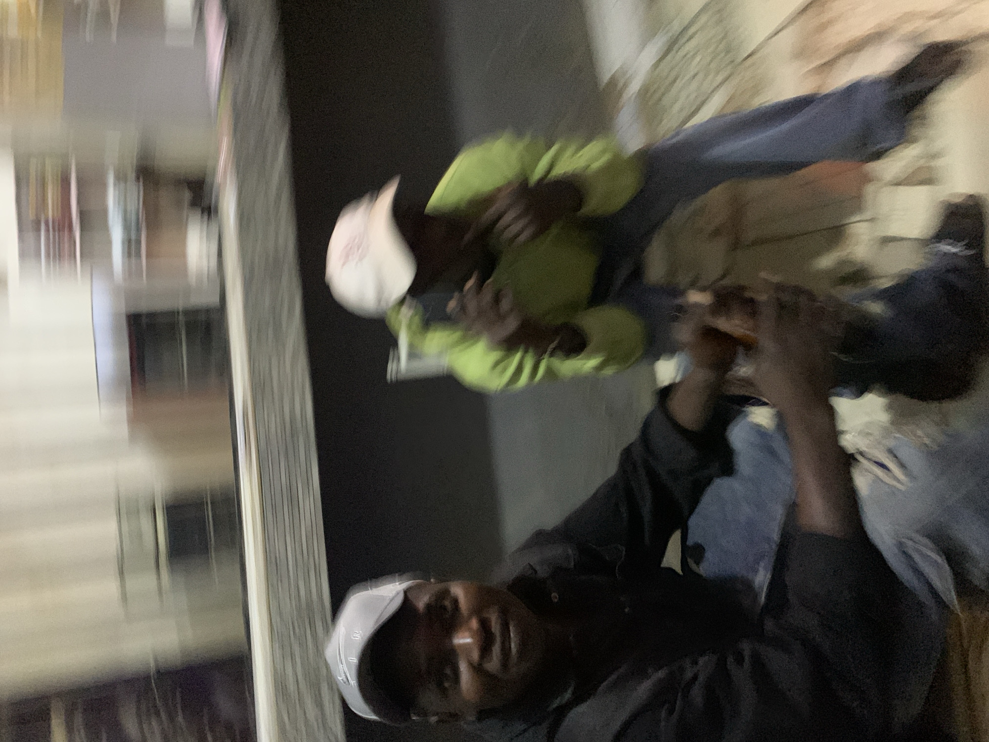 This image captures two adult males seated in a dimly lit indoor environment within the city of New Stadium, Nigeria. The photograph is characterized by significant motion blur, particularly affecting the background and the individual on the right.
The person on the left, an adult male of African descent, is seated and directly facing the camera with a subtle smile. He wears a dark, long-sleeved shirt or jacket and light blue jeans, which appear to have distressed or ripped details at the knees. A grey baseball cap, featuring a distinct white "R" emblem on the front, is worn on his head. His hands are clasped around an indistinct, elongated object, possibly a bottle or can, which rests between his legs.
To the right and slightly behind the first individual, a second adult male, also of African descent, is seated. He is dressed in a bright lime green or yellow-green hooded sweatshirt and dark blue pants. A white baseball cap covers his head. His facial features are heavily obscured by blur, and his head appears to be angled away from the camera's direct view. His hands are raised in front of him, in an ambiguous gesture or holding an unidentifiable object due to the severe blur.
The background is extensively blurred, showing horizontal streaking consistent with motion. It suggests an interior setting, possibly a retail space or a room with shelving units. Light-colored walls are discernible, along with shelves containing various indistinct items, some appearing reddish-brown and others bluish. A dark horizontal surface, resembling a counter or table, spans the mid-ground of the scene. The overall lighting is low, indicating a potential evening or night setting, or an interior space with limited artificial illumination.