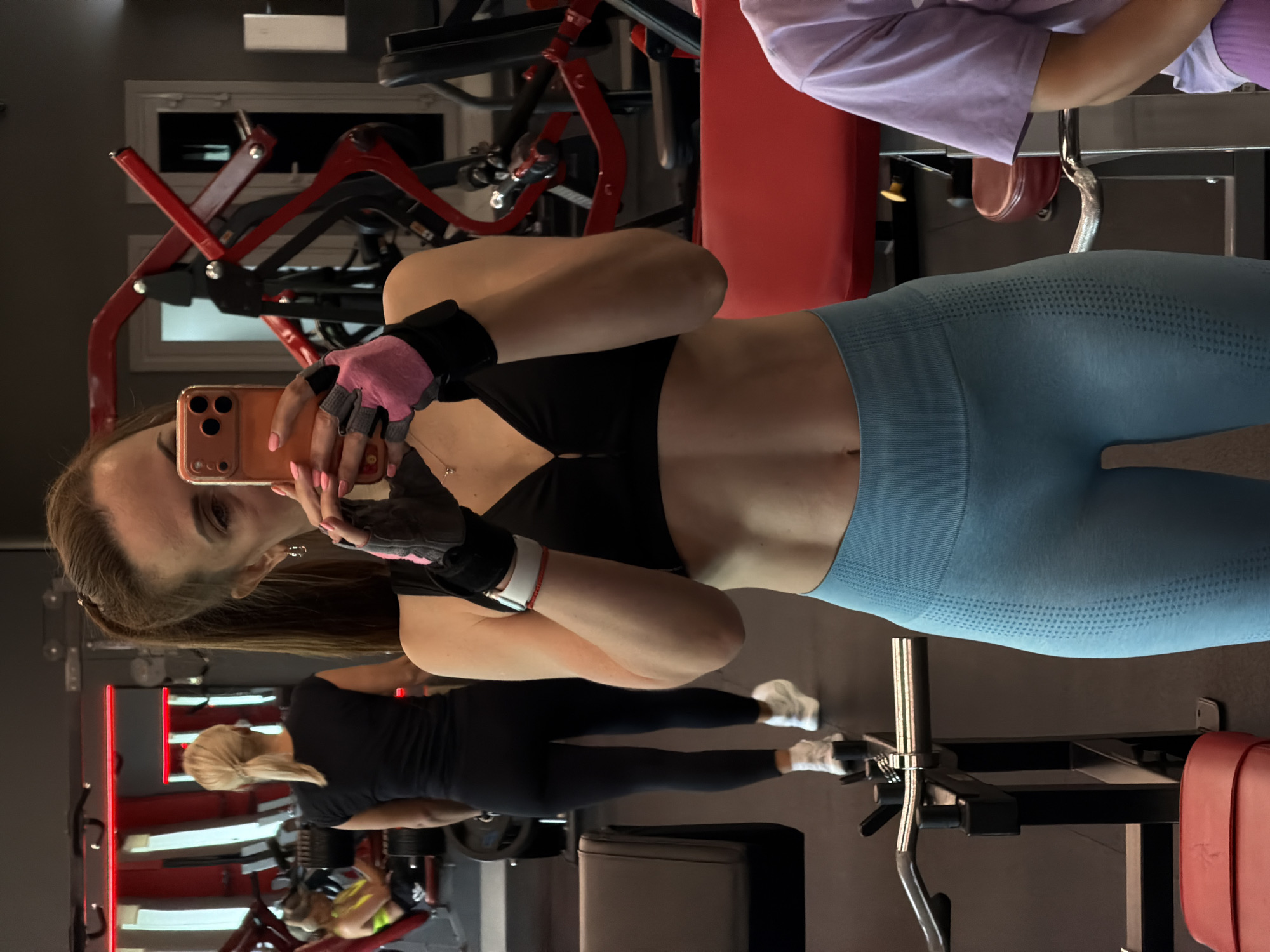 A young woman is taking a selfie in a gym in Birobidzhan, Russia. She is wearing a black sports bra, blue leggings, and pink and grey gym gloves. Her hair is pulled back in a ponytail. In the background, another woman in a black outfit is walking away from the camera. The gym appears to be well-equipped with various weight machines and equipment. The lighting suggests an indoor setting.