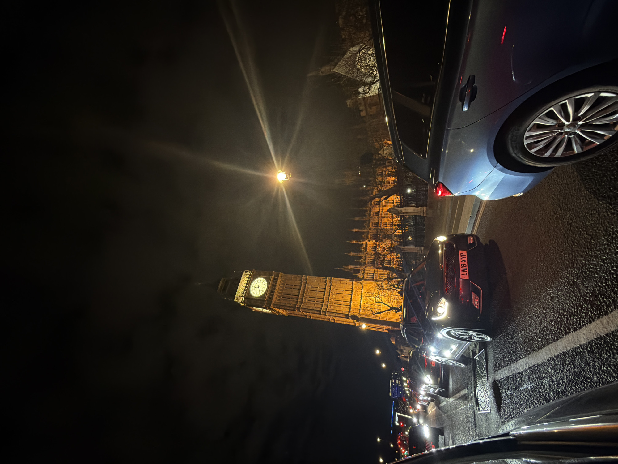 At night, the Houses of Parliament and Elizabeth Tower (Big Ben) are illuminated, with the clock face clearly visible. Cars are lined up on a street in front of the landmark. A blue car is positioned on the right edge of the frame, while a black car with the license plate "LN18 XYP" is centered in the foreground. Other vehicles are also visible in a line behind the black car. Streetlights illuminate the scene, and the sky is dark. The environment appears to be in the city of London, United Kingdom.