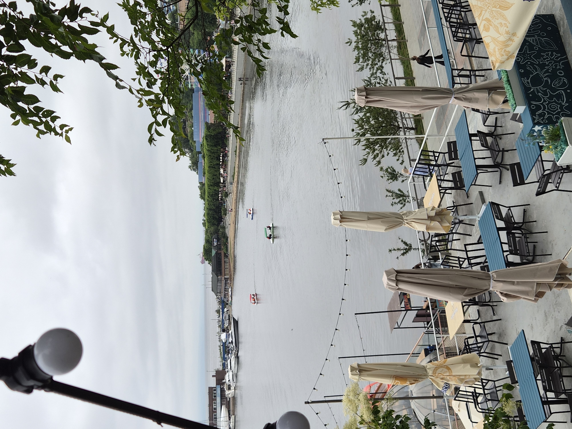 The image shows an outdoor seating area of a restaurant or cafe overlooking a harbor in Vladivostok, Russia. The foreground features tables and chairs, many covered with umbrellas. In the middle ground, a body of water is visible, with several small boats and pedal boats on it. Along the far bank of the water, there is a promenade with people walking, as well as buildings and trees. In the distance, more boats are docked at a marina, and the sea stretches to the horizon under a cloudy sky. The overall scene depicts a waterfront recreational area.