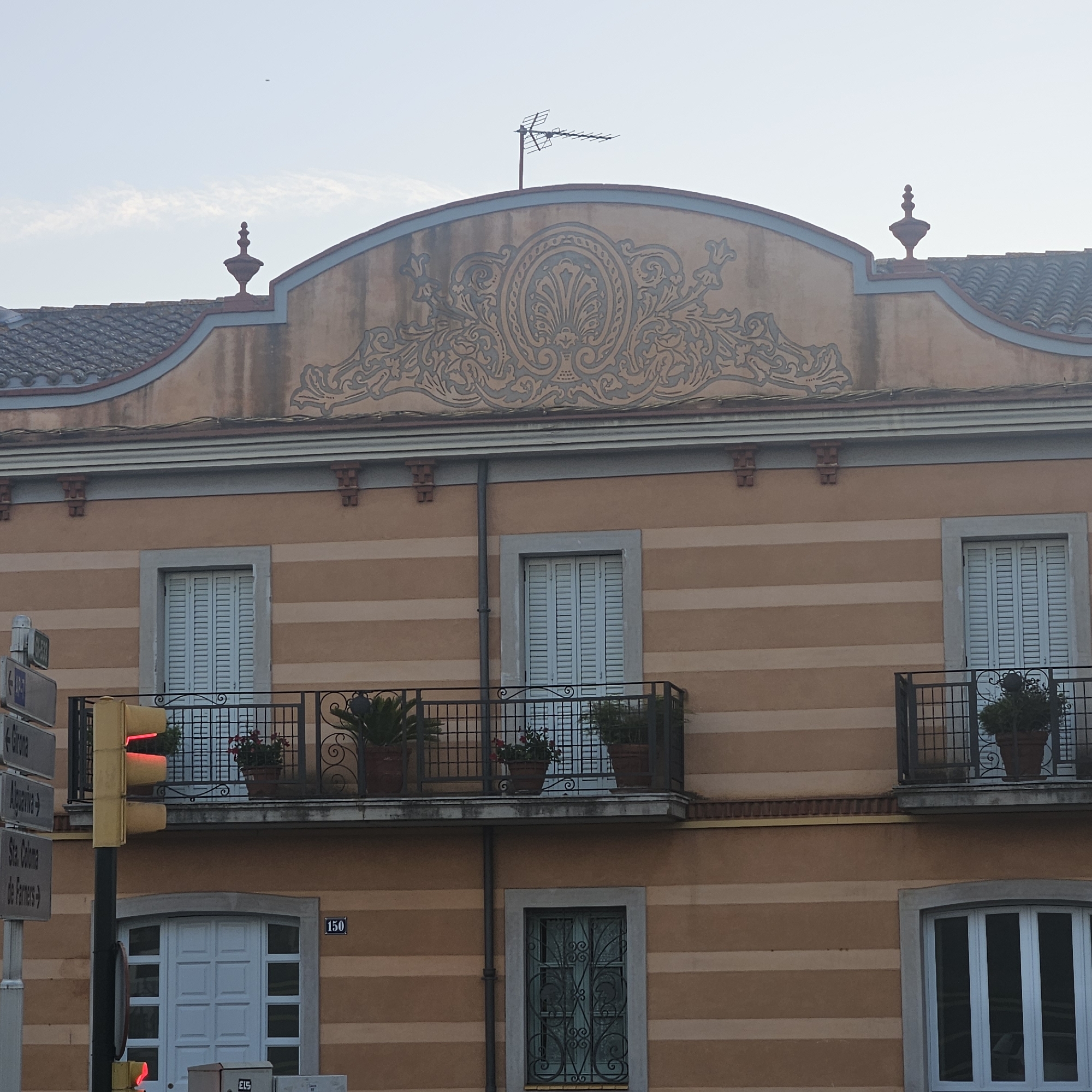 The image shows the facade of a building in El Perelló, Spain. The building is painted in horizontal stripes of a peachy orange color. The upper part of the building features a decorative frieze with a central ornamental motif. There are two balconies with black wrought-iron railings and potted plants. Below the balconies, there are several windows, some with light blue shutters and others with ornate metal grilles. A traffic light with a red light illuminated is visible on the left side of the image, along with street signs. A small black object, possibly a drone or a bird, is in the sky.