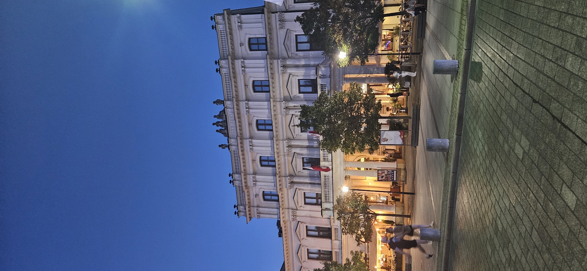 The image shows the front of a large, ornate, neoclassical building at dusk. The building features multiple stories with numerous windows and decorative columns. Two flags, one red and white, are visible on balconies. The sky is a deep blue, indicating twilight. In front of the building, there is a street with people walking and sitting at outdoor cafe tables. Trees line the sidewalk. The ground in the foreground is paved with cobblestones. Streetlights are illuminated. One can infer this is likely a public square or a main street in a European city given the architectural style and the presence of outdoor seating. Based on the architecture and context, this appears to be the Teatr Wielki – Polish National Opera in Warsaw, Poland.