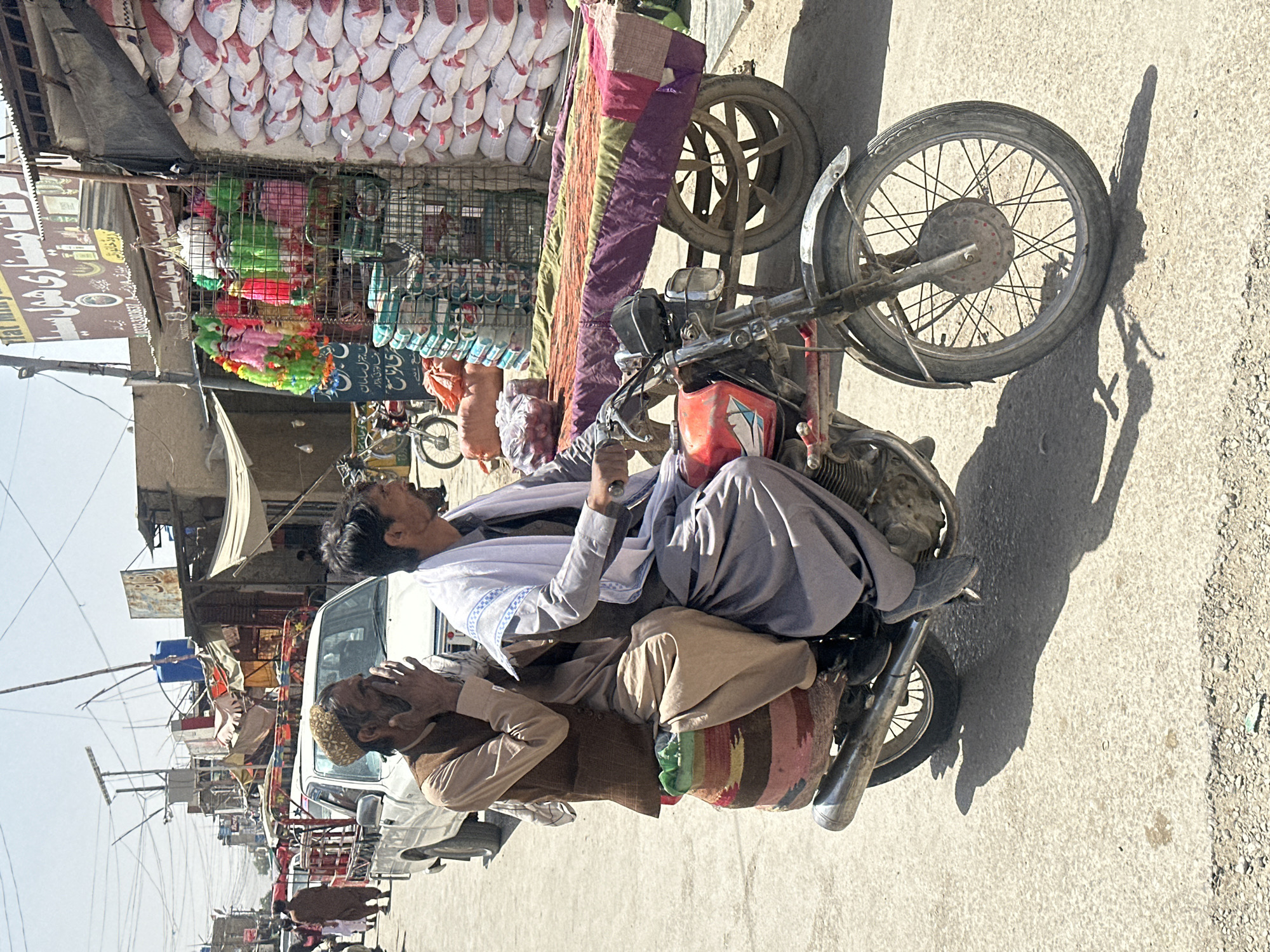 In a bustling street scene in Kanozai, Pakistan, two men are riding a red motorcycle. The man in the front, the driver, is wearing a grey outfit and a white scarf draped around his neck. The man behind him, a passenger, is dressed in a brown vest over a tan shirt and trousers, with his right hand covering his mouth. He wears a traditional embroidered cap. Both men appear to be engaged in conversation or reacting to something. They are riding past a market stall laden with large sacks, possibly of grain, stacked high. To the left of the stall, colorful garlands and other items are displayed. A white pickup truck is visible further down the street, along with other pedestrians and parked bicycles, indicating a busy commercial area. The environment is characterized by a dusty, sun-drenched street and simple, functional market structures.