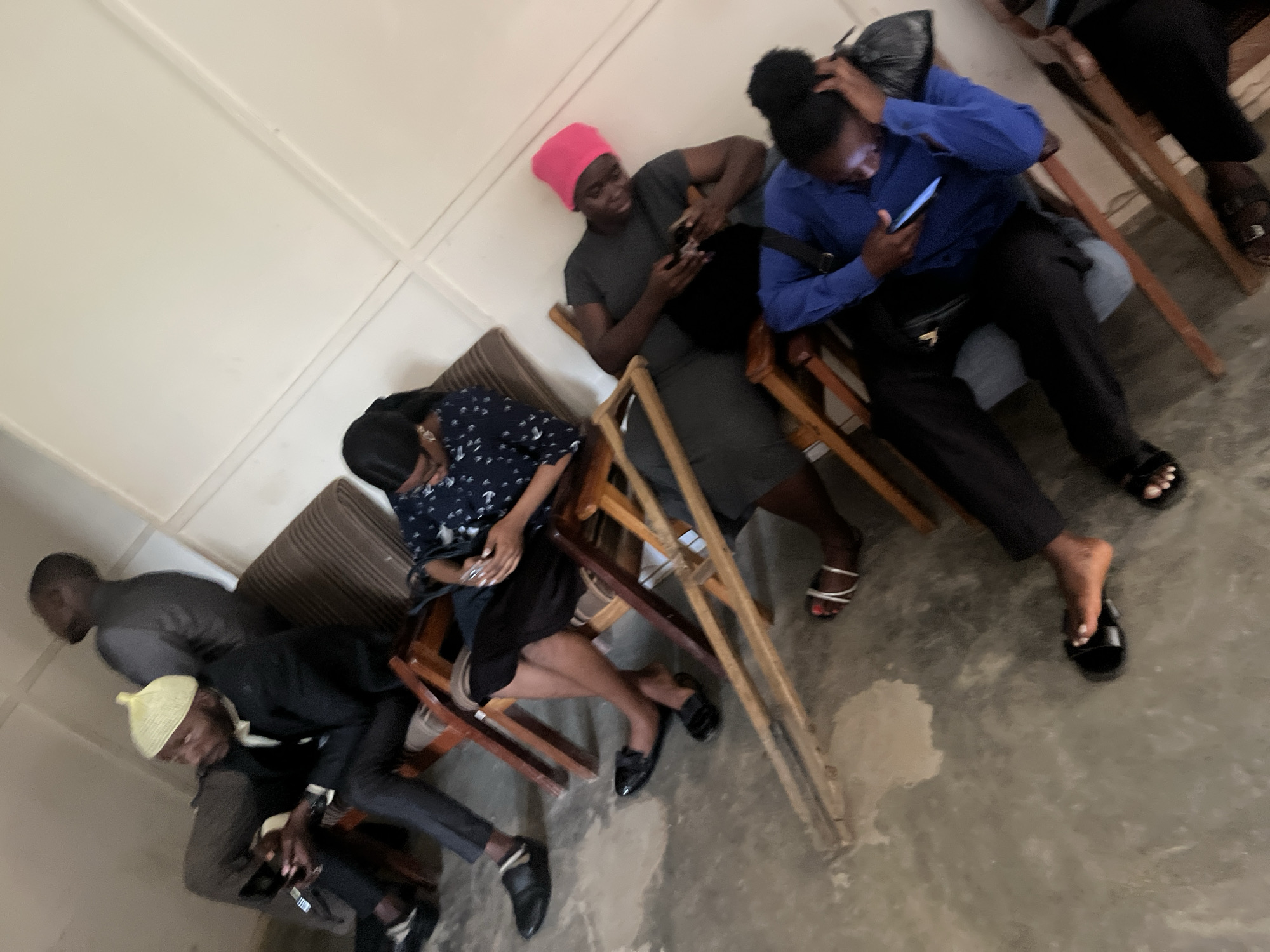 The image depicts a group of people, likely in a waiting area or communal space, in Jos, Nigeria. The scene is indoors, with off-white walls and a concrete floor.
In the foreground, a person in a blue long-sleeved shirt and black pants is seated, engrossed in their phone. To their left, another individual in a dark grey t-shirt and black pants, wearing a bright pink head covering, is also looking at their phone. Behind them, a woman in a dark patterned blouse and black skirt is seated and appears to be using her phone.
Further back, a man in a black suit and a yellow cap is seated, holding a phone in his hand. Next to him, another man in a dark suit is standing. The seating arrangement consists of simple wooden chairs with fabric cushions. Some of the chairs appear to be in a state of disrepair, with visible wooden supports. There are also some wooden planks on the floor in the middle of the image, possibly part of damaged furniture or construction. The overall impression is one of a functional, but not particularly well-maintained, indoor public space.