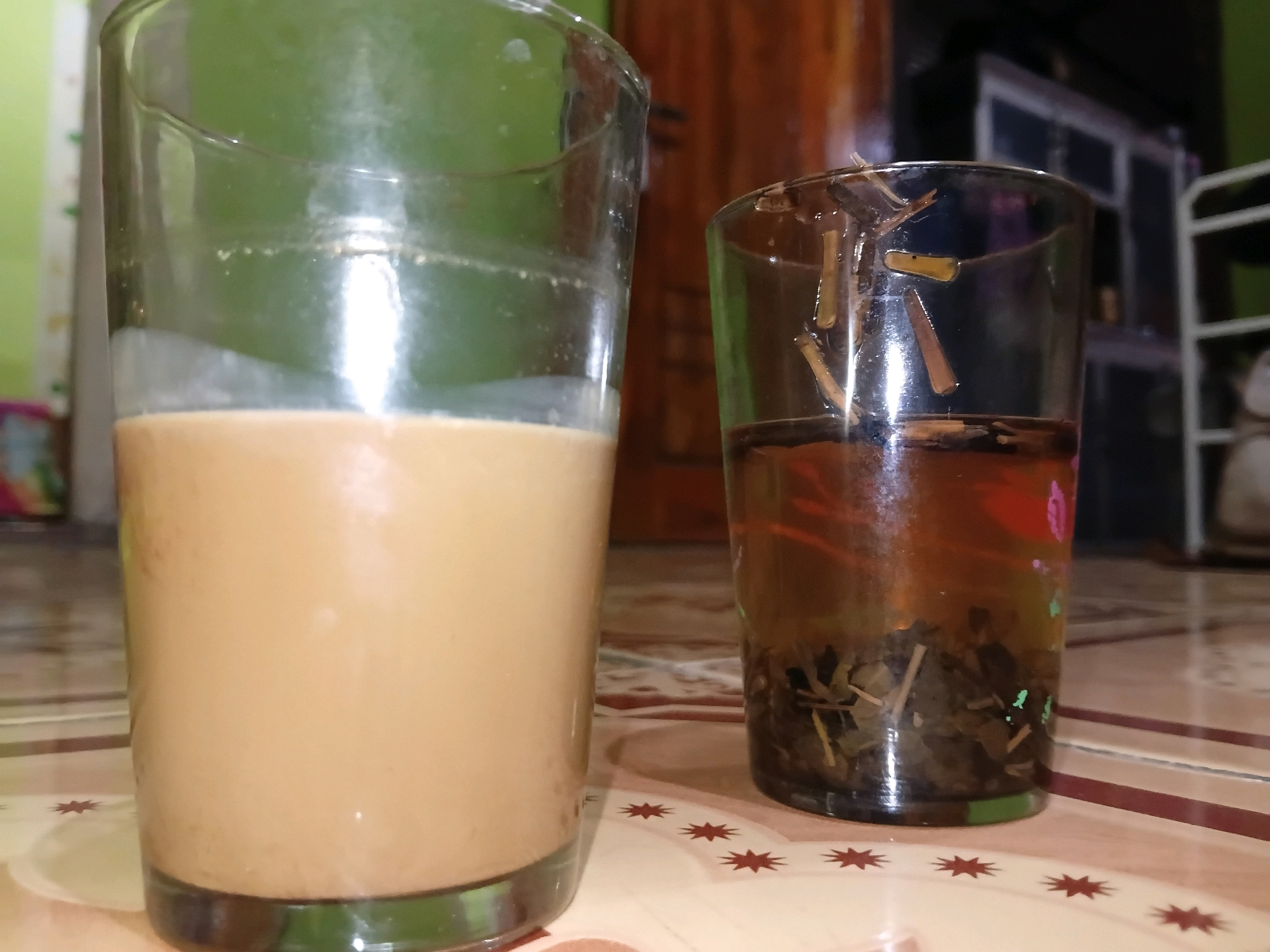 Two glasses of beverages are placed on a patterned surface. The glass on the left contains a light brown liquid, likely coffee or tea with milk. The glass on the right contains a darker amber liquid with visible tea leaves at the bottom and some floating pieces on top. The background is blurred, showing a green wall, wooden furniture, and shelves with some items. The scene appears to be indoors. The location is Margo Rukun, Indonesia.