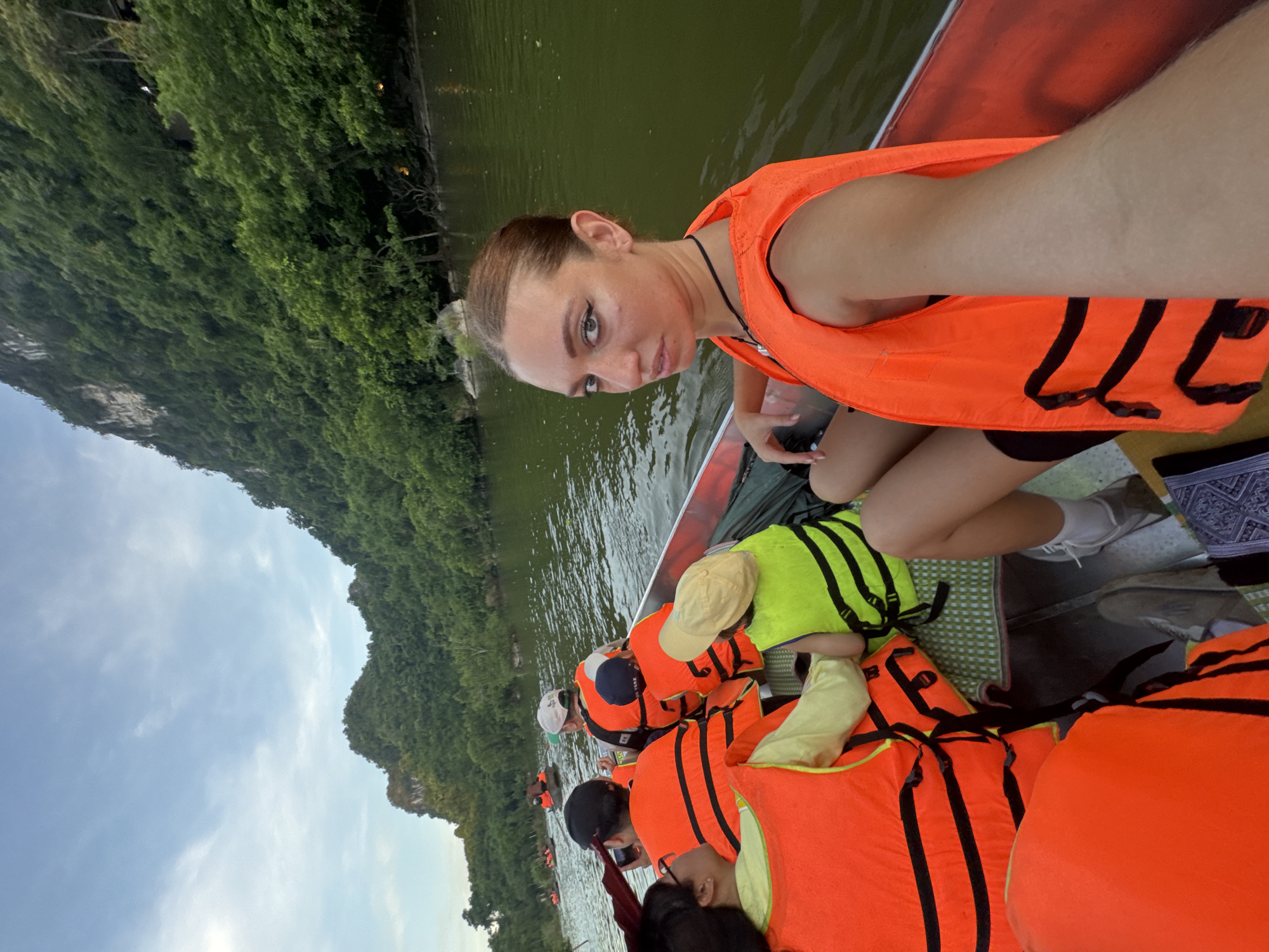 In the city of Yên Sơn Ward, Vietnam, a group of people are on a boat excursion. The photo features a young woman in the foreground, wearing an orange life vest and looking at the camera. She is in a boat with several other individuals, also wearing life vests, suggesting a recreational activity on the water. The background displays a lush, green landscape with steep, tree-covered hillsides and a calm body of water. The sky is partially cloudy, indicating either early morning or late afternoon. The presence of multiple boats in the distance suggests this is a popular tourist area.