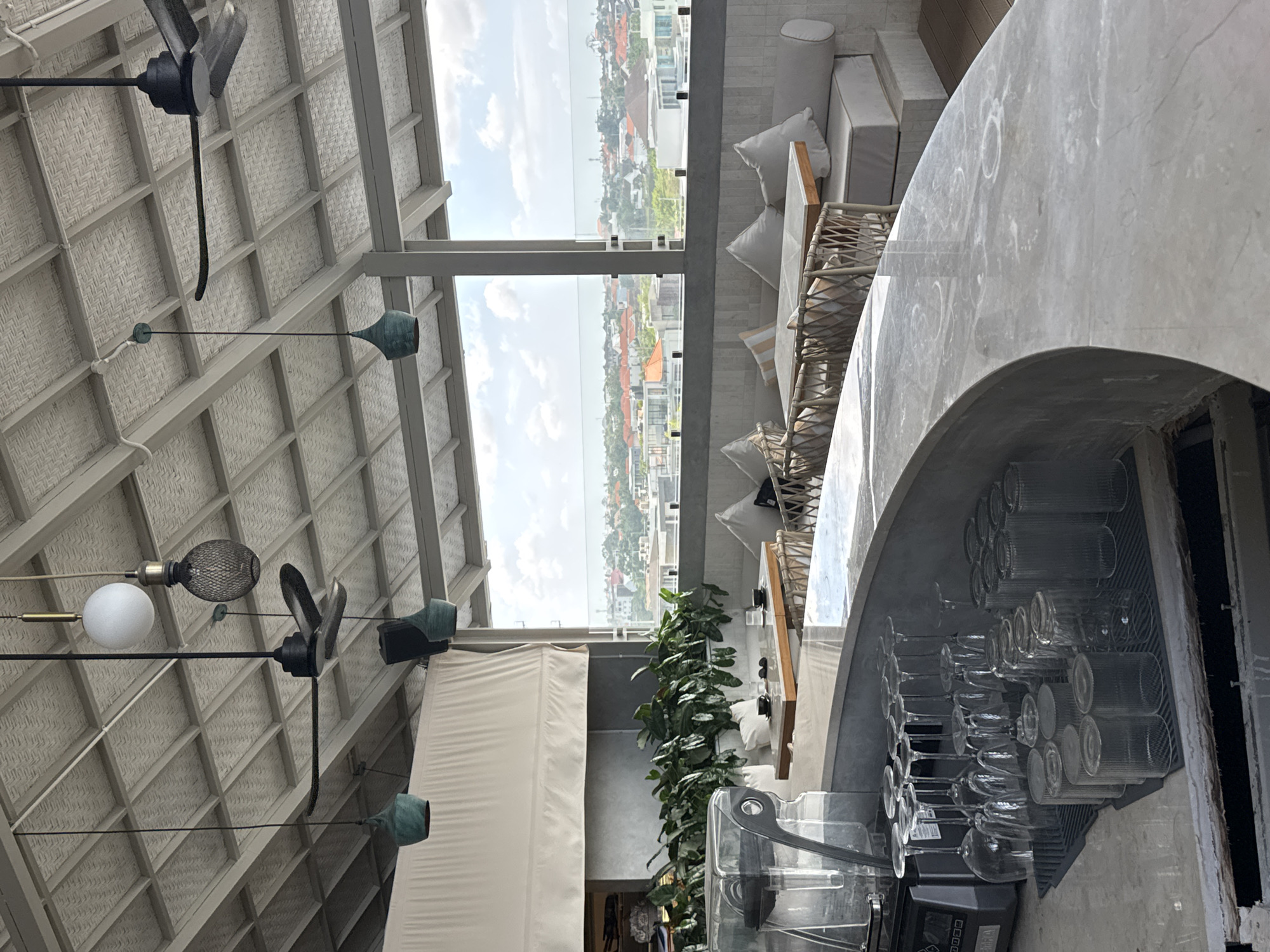 The image shows an interior view of a rooftop bar or restaurant in North Kuta, Indonesia, with a view of the city in the background. In the foreground, a marble-topped bar is visible, with stacks of clean wine glasses and tumblers arranged on a gray mat. A blender or coffee machine is also present on the bar. The bar area appears to be outdoors or semi-outdoors, with a sloped ceiling made of textured panels and visible beams. Several industrial-style ceiling fans and hanging pendant lights are attached to the ceiling.
Behind the bar, a seating area with tables and chairs is set up. The tables are made of wood and marble, and are surrounded by woven wicker chairs. Cushioned benches with white and striped pillows line the walls. Large potted plants with dark green leaves are placed behind the seating area, adding a touch of greenery.
The background features a panoramic view of North Kuta, with a dense arrangement of buildings, many with terracotta-tiled roofs, stretching towards the horizon. The sky is partly cloudy. The overall ambiance is modern and airy, designed for enjoying the city view and tropical climate. No people are visible in the image.