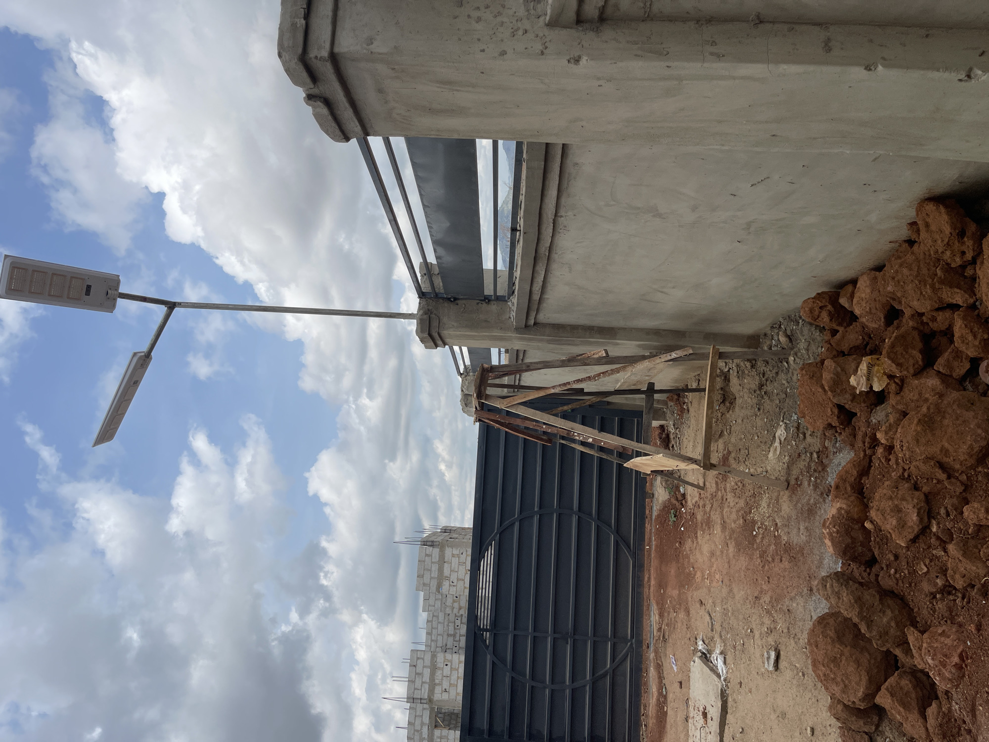 The image depicts an outdoor scene in Angwan Sayawa, Nigeria, showing construction. In the foreground, there is a black metal gate and wooden scaffolding. A pile of reddish-brown rocks is adjacent to the gate and scaffolding. Behind the gate, there is a partially constructed structure of grey cinder blocks. Above the structure, the sky is visible with a mix of blue and white clouds. A street light is visible. A grey concrete structure is in the background.