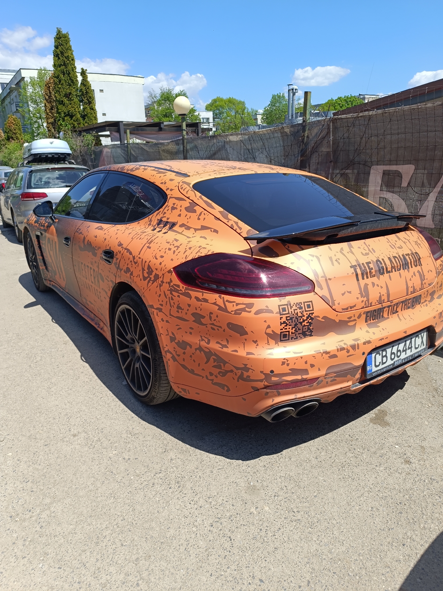 Here is a description of the image:
The image shows an orange Porsche Panamera parked on a city street in Sofia, Bulgaria. The car has a distinctive custom wrap featuring a camouflage pattern and lettering that reads "THE GLADIATOR: FIGHT TILL THE END." A QR code is also printed on the rear. The car's license plate reads "CB 6644 CX," which is a Bulgarian license plate.
The car is parked on an asphalt street in a parking spot. Other cars are parked nearby. A fence is visible in the background, with greenery and buildings behind it. The sky is blue with some clouds. It is a sunny day.