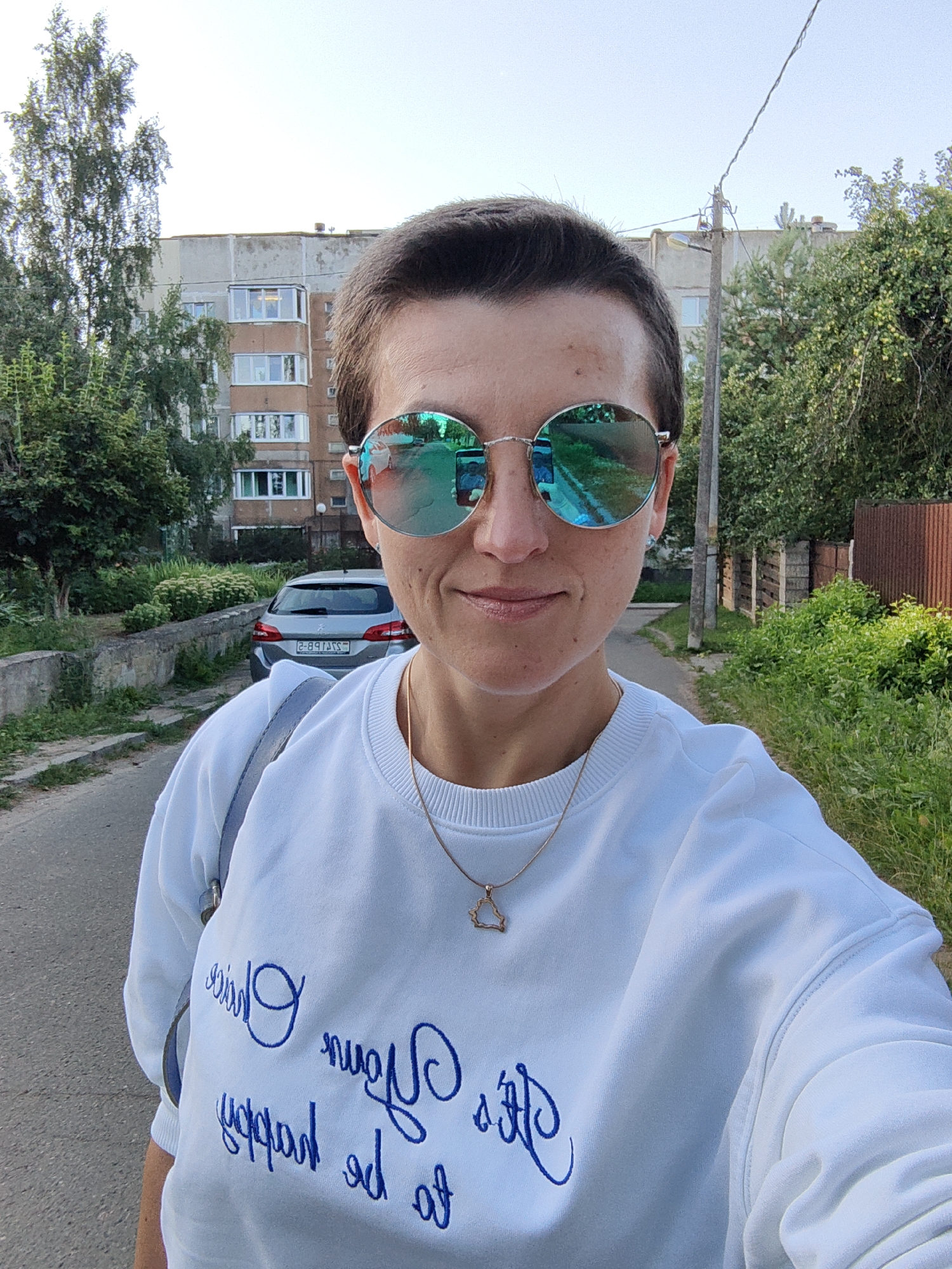 A woman with short, dark hair and sunglasses with green mirrored lenses is taking a selfie outdoors. She is wearing a white sweatshirt with blue cursive writing on it and a thin gold necklace with a triangular pendant. Behind her, a grey car is parked on a paved road lined with trees and a multi-story apartment building. The scene appears to be in a residential area of Fanipal, Belarus, given the architectural style of the building and the license plate of the car. The sun is shining, creating shadows and highlighting the green foliage.