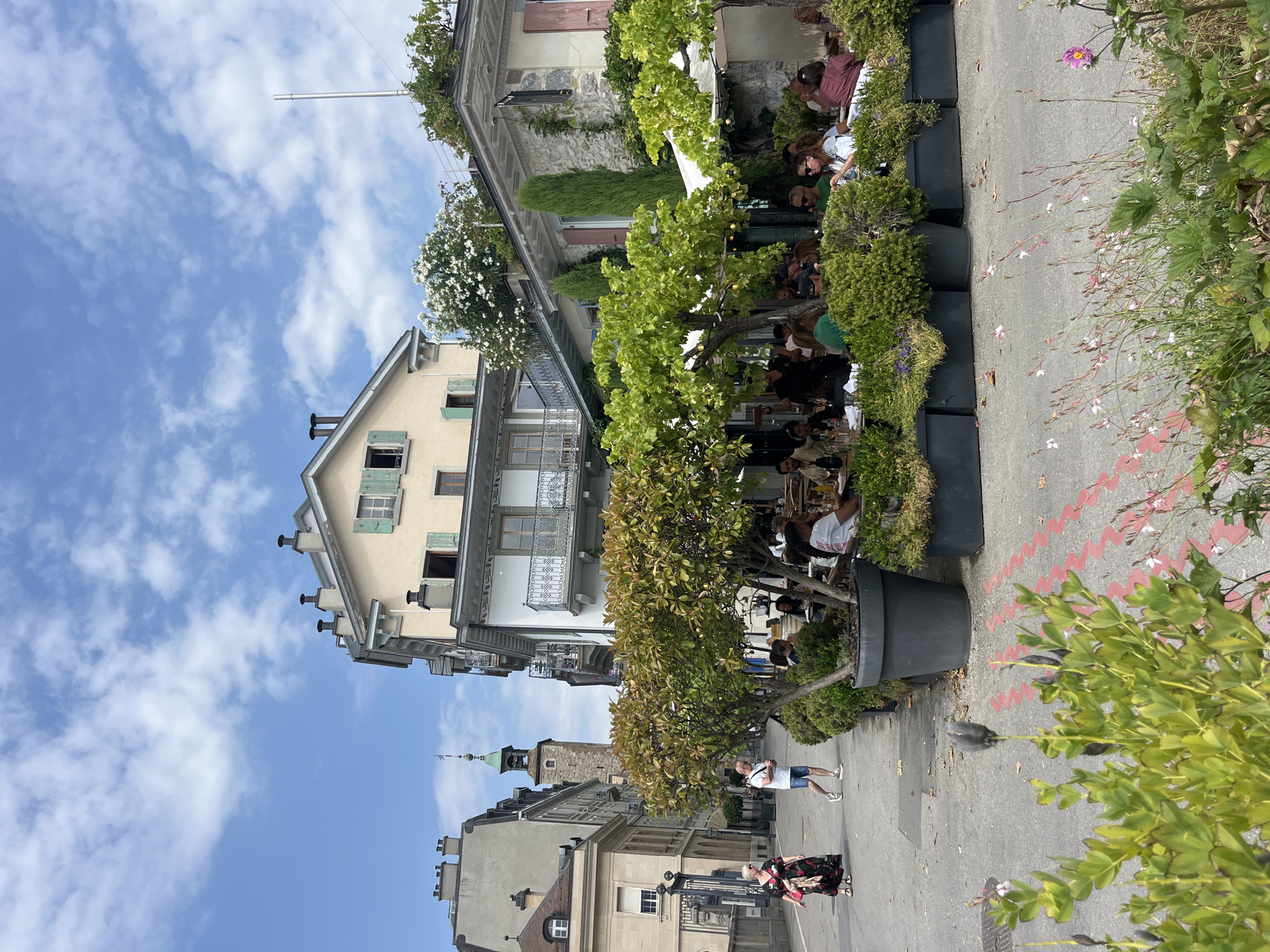 The image depicts a street scene in Vevey, Switzerland, likely during the daytime, under a partly cloudy sky. In the foreground, a sidewalk is visible with some greenery and flowers. A woman in a patterned dress is walking on the sidewalk to the left, followed by a man in a t-shirt and shorts. To the right, an outdoor seating area for a restaurant or cafe is set up, with tables and chairs occupied by people dining. Large planters filled with trees and shrubs line the seating area, separating it from the sidewalk. In the background, buildings of varying architectural styles are visible, including one with a distinctive tower. The atmosphere suggests a leisurely day in a European town.