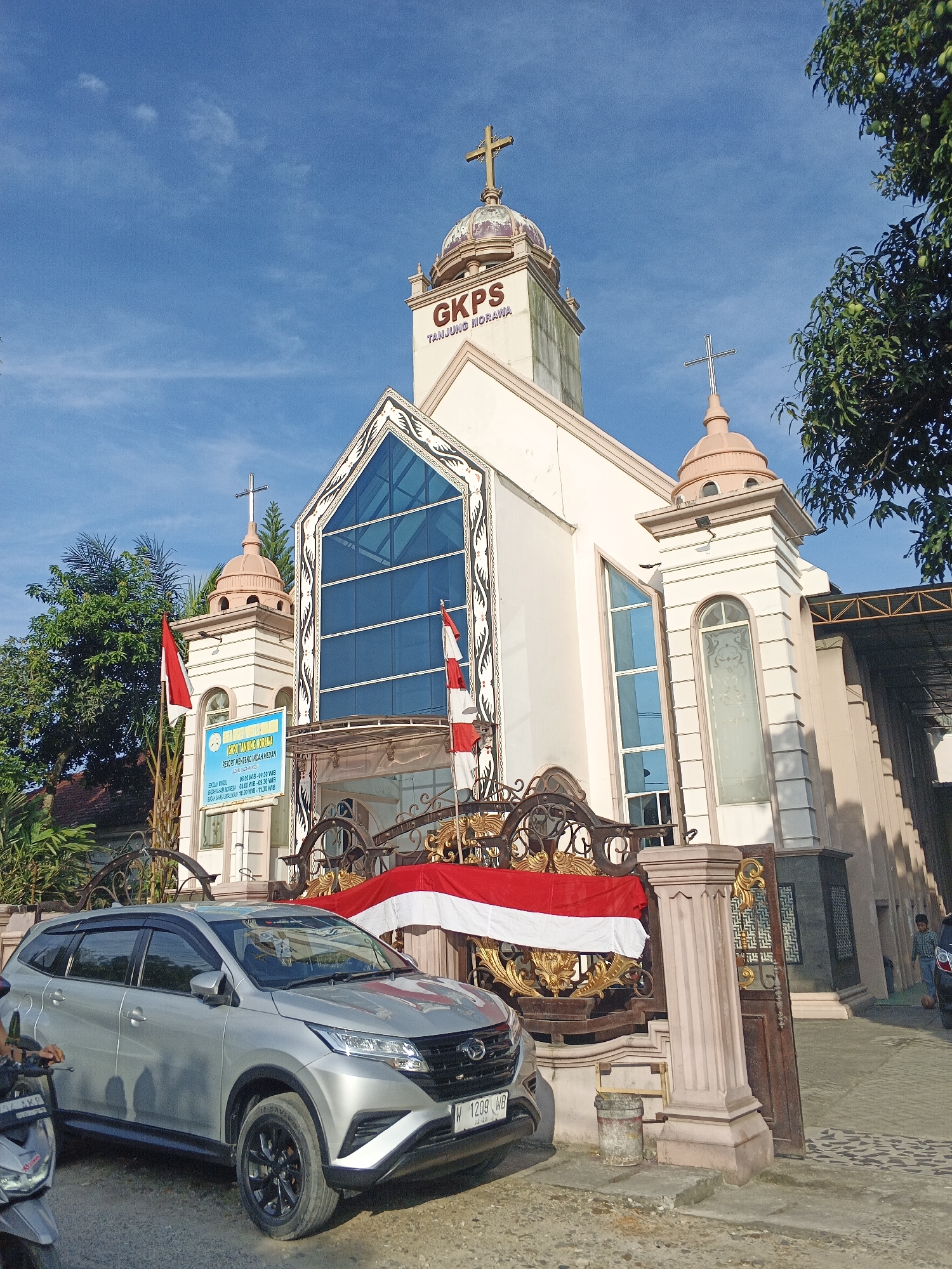 The image shows the exterior of a church in Limau Manis, Indonesia, identified by the signage on the building as "GKPS TANJUNG MORAWA". The church is a light-colored building with multiple towers topped with crosses. One prominent tower has the church's name in large red letters. The facade features large blue-tinted windows and decorative elements. In front of the church, a silver SUV is parked on a paved area, with a motorcycle partially visible on the left. A decorative metal fence, adorned with gold accents, stands in front of the church, and two Indonesian flags (red and white) are displayed on poles and draped over the fence. The sky is blue with some scattered clouds. Trees with green foliage are visible on the right and left sides of the image, and part of a mango tree is visible in the upper right. The overall scene depicts a well-maintained religious building in a somewhat tropical environment.