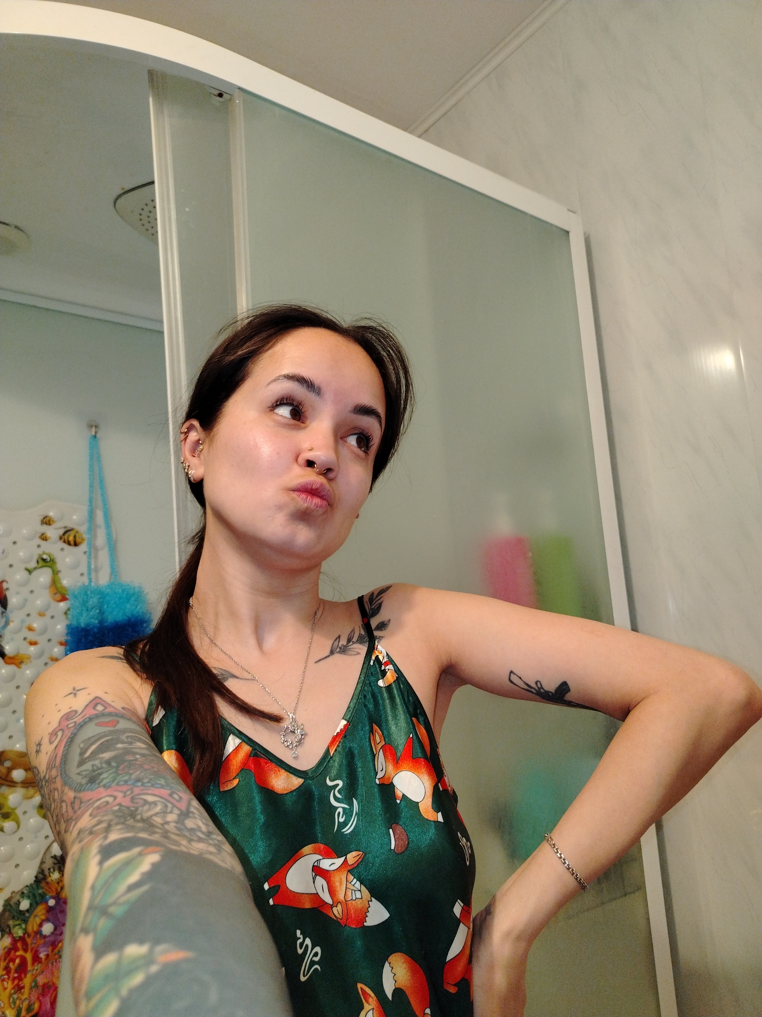 A woman is posing in a bathroom, likely in a domestic setting, as inferred by the presence of a shower and toiletries. The woman has dark hair pulled back in a ponytail, and multiple piercings in her left ear. She is wearing a green top with a fox pattern and a silver necklace. She has tattoos on both arms, including one depicting a weapon on her right arm. She is making a pouting expression. The background includes a shower stall with frosted glass, a shower head, and a tiled wall. There are also some items hanging in the shower. The location is not possible to confirm because the image does not provide specific geographic details related to the city of Noviy, Russia.