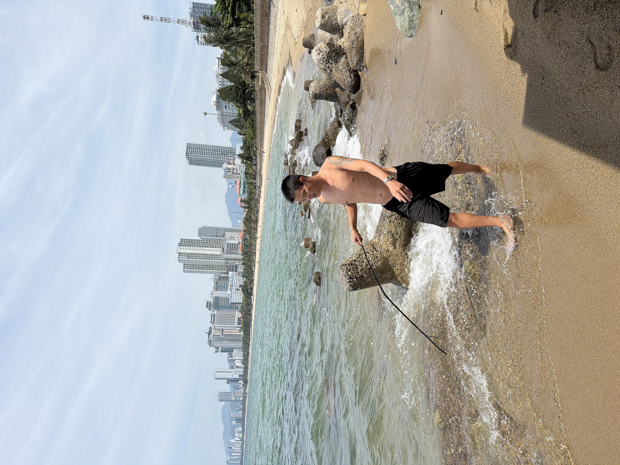 A shirtless man in black shorts walks along the shoreline in Nha Trang Ward, Vietnam, holding a stick and wading through shallow water. The beach is lined with concrete tetrapods and rocks. In the background, a modern city skyline with numerous high-rise buildings stretches along the coast under a bright, cloudy sky. The ocean exhibits clear, greenish water with gentle waves lapping the sandy shore.