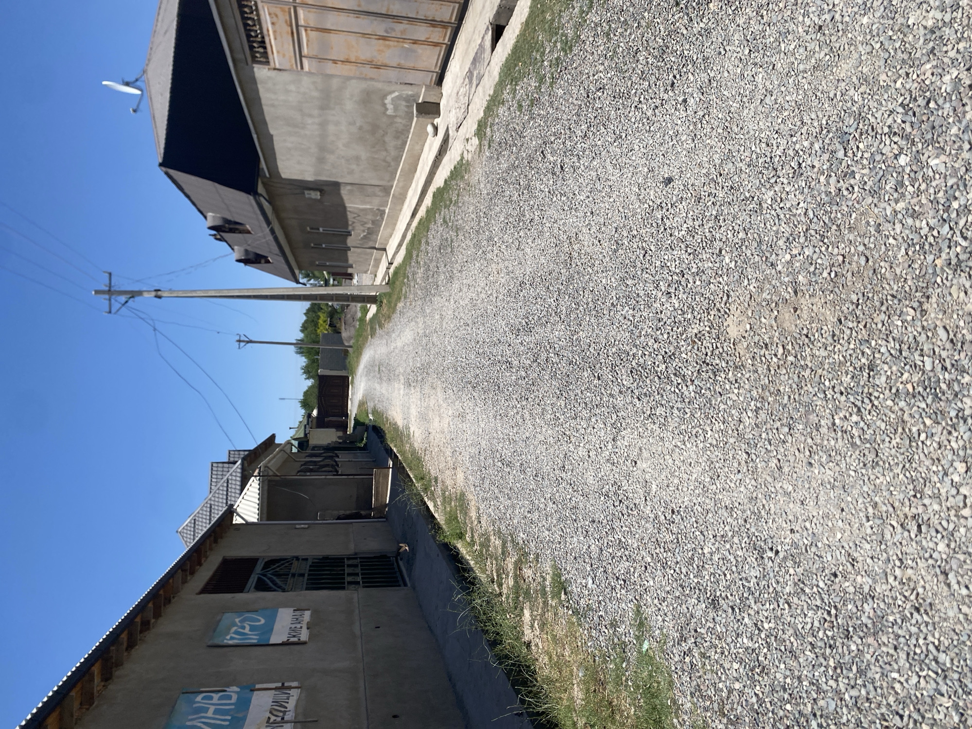 The image shows a gravel road leading into a village in Bogiston, Tajikistan. On the left, a building with a gray facade has blue signs with white text, and a metal gate. Further down the road, there are more buildings with similar gray facades, some with tiled roofs, and a large, rusted metal gate. On the right, another building with a gray facade and a dark tiled roof features a satellite dish. A utility pole with wires stands on the right side of the road, and trees are visible in the background. The sky is clear and blue.