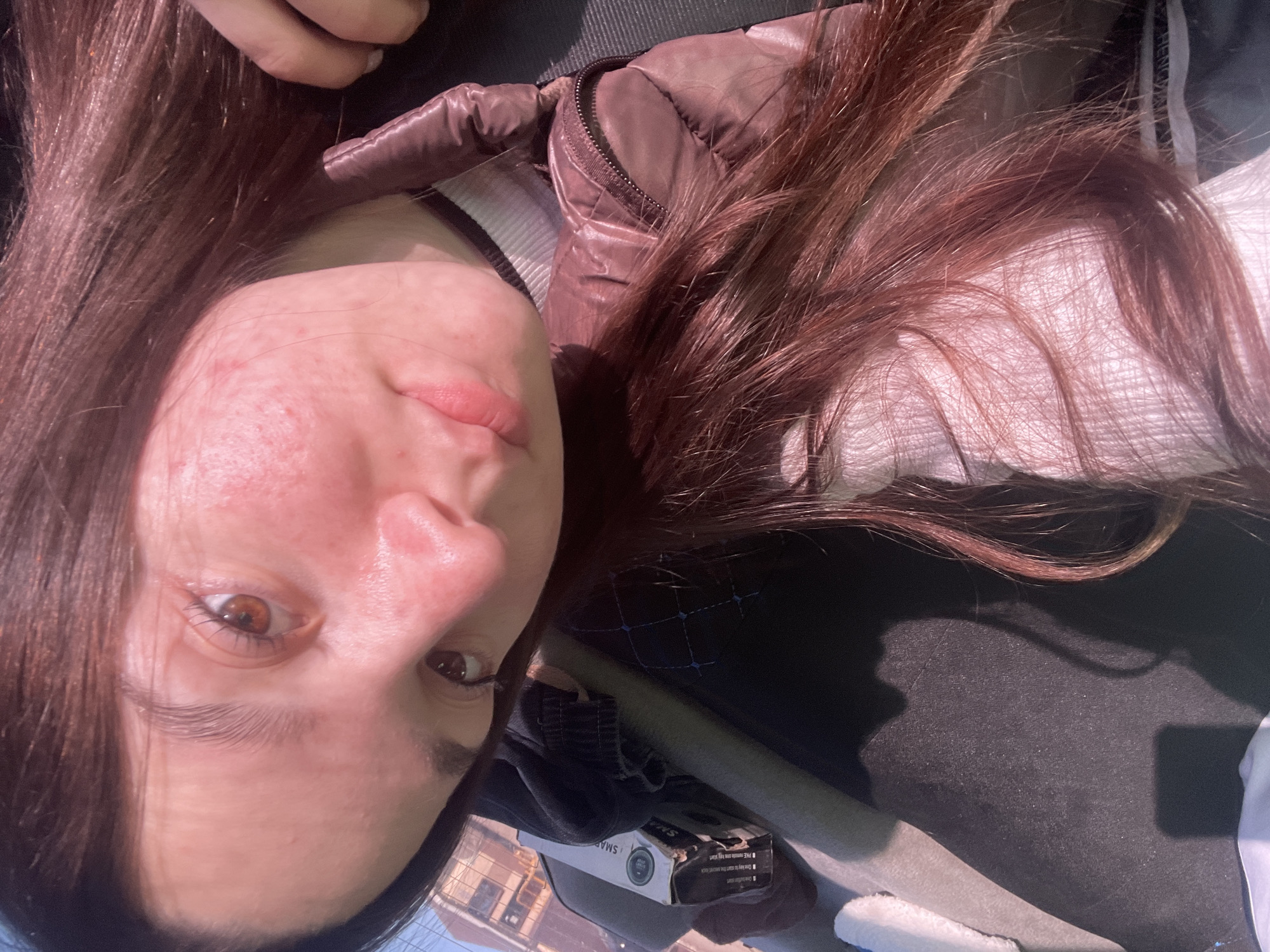 A rotated selfie of a woman with long brown hair and light skin is centered in the frame. Her right cheek, closest to the camera, shows some skin imperfections. She has brown eyes and a neutral facial expression. The woman wears a white top with a dark gray and white striped collar, and a brown puffy jacket partially visible on the left and top right of the image. The background is a mix of dark surfaces, potentially a vehicle's interior and black laptop. A product box with the words "Smart Camera" is situated below the woman. Outside the car window is visible architecture, indicating a building with windows, suggesting an urban setting. Location, Chaikovsky, Russia, is not confirmed.
