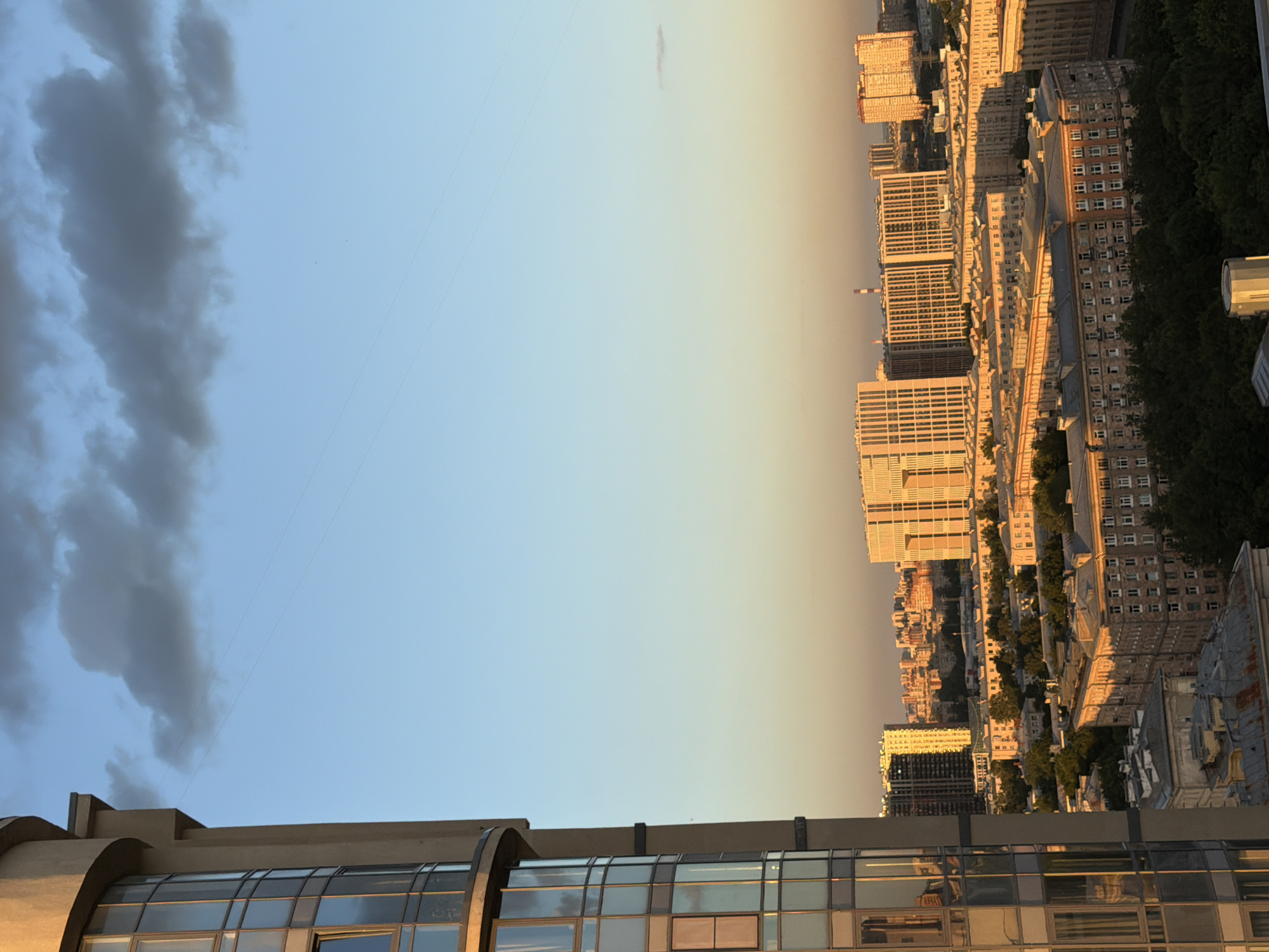 An exterior, high-angle, vertical medium shot captures a cityscape at sunset. On the left, a curved, modern building with numerous glass windows dominates the foreground. To the right, an expanse of sky transitions from a clear, light blue at the top to a warmer, golden hue near the horizon. A single, dark cloud is visible in the upper sky. Below the sky, the urban landscape unfolds with numerous buildings of varying architectural styles and heights. In the midground, several large, modern buildings with repetitive window patterns are illuminated by the setting sun, casting a warm glow. These are surrounded by lower, older buildings with more traditional facades, many featuring multiple stories and smaller windows. Dense greenery, likely trees, can be seen interspersed among the buildings in the lower part of the cityscape. A faint wire is visible stretched across the sky. The scene depicts a densely populated urban environment bathed in the soft light of evening.