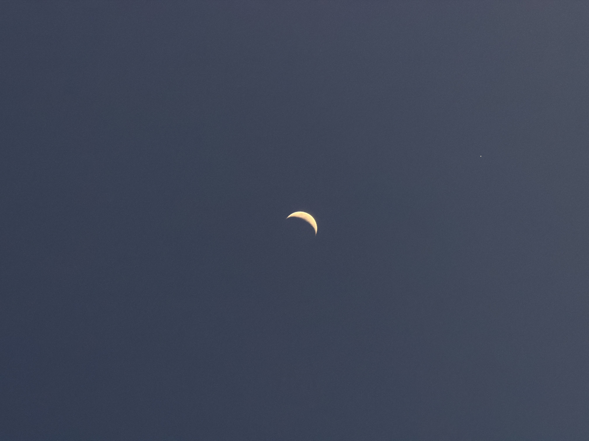 The image shows a crescent moon in a dark blue sky. To the right of the moon is a small, bright point of light, possibly a star or planet. The environment is a night sky. No specific location in Moscow, Russia, can be determined from the image. No people, actions, or interactions are depicted.