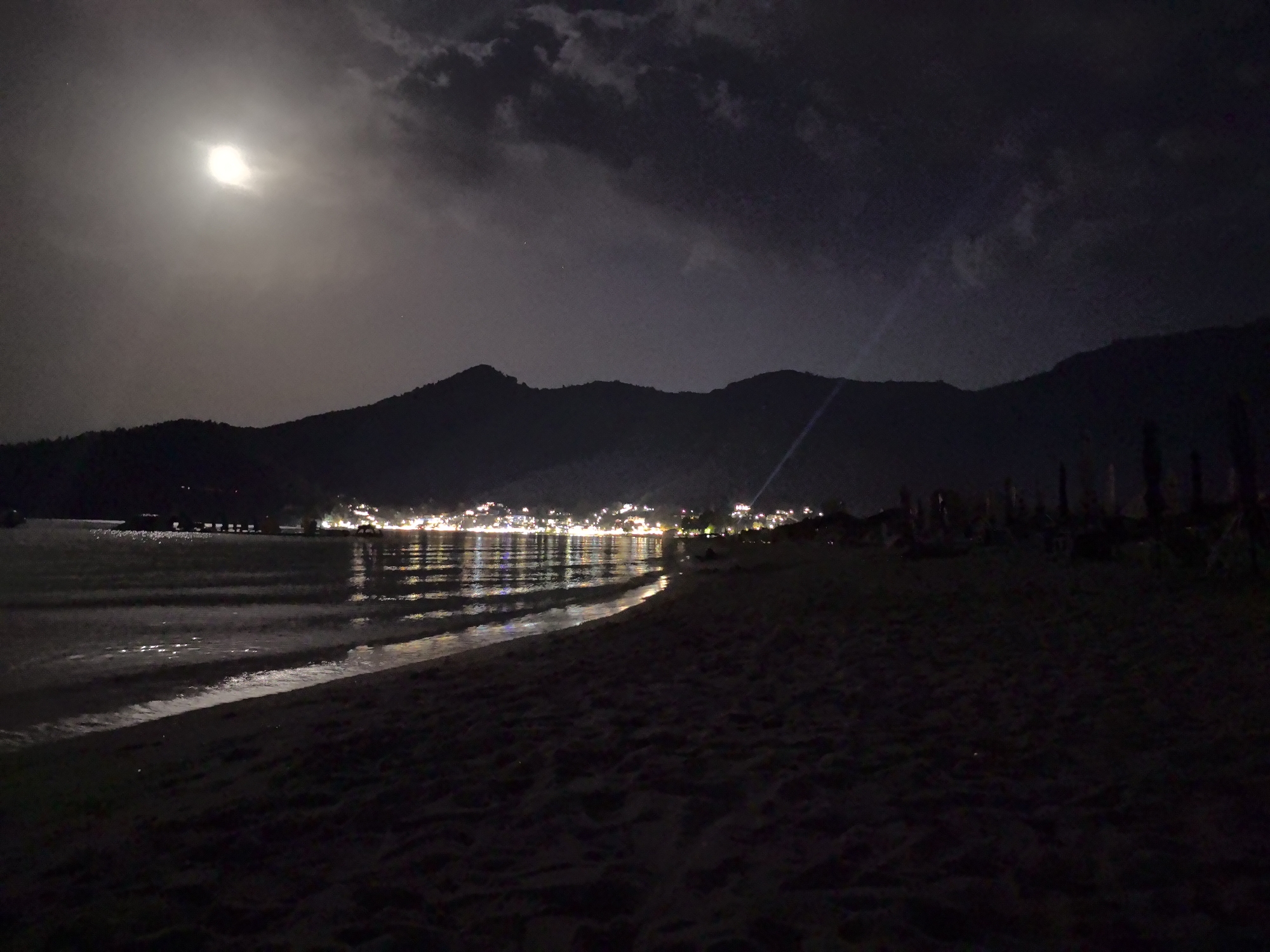 At night on a beach in Golden Beach, Greece, the moon is visible in the sky above a silhouette of mountains. A town with lights reflecting on the water is seen in the distance. The beach is dark, with some beach chairs and umbrellas visible. The sea is calm with small waves lapping at the shore.
