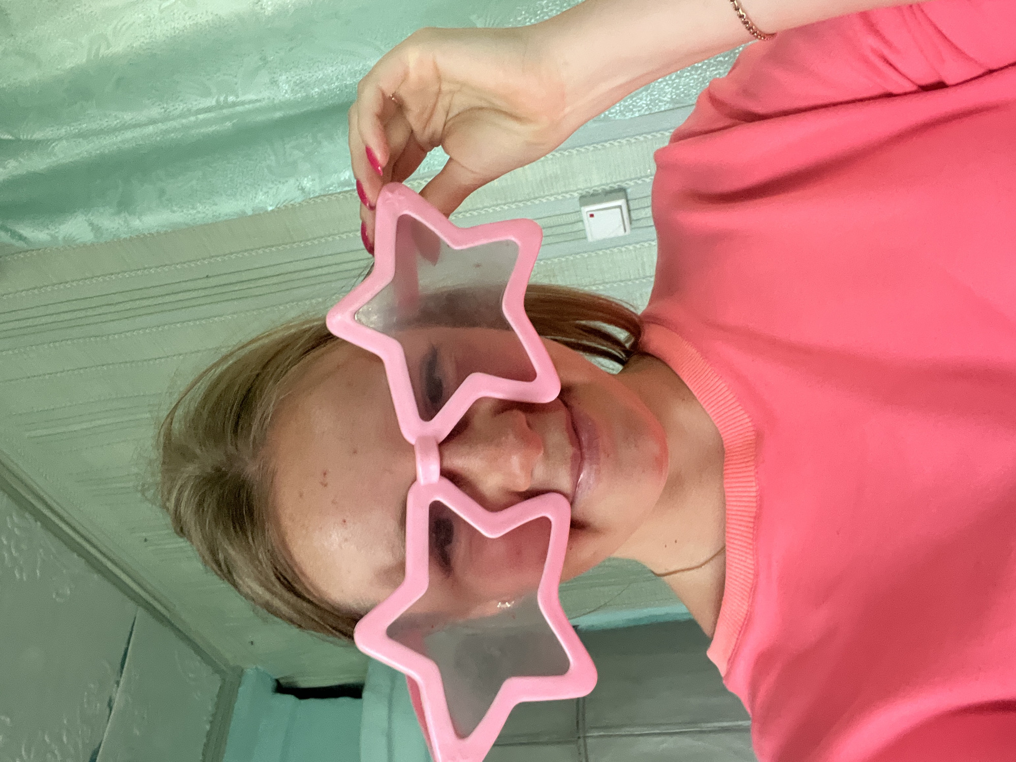 The image shows a close-up of a person wearing large, pink star-shaped novelty glasses. The person's face is visible, with their eyes looking through the star-shaped lenses. They are wearing a bright pink top. The background appears to be a room with light green patterned wallpaper and horizontal lines, possibly suggesting a ceiling or a wall. A white light switch is visible on the wall. The lighting suggests it is daytime. The overall mood is playful and lighthearted due to the novelty glasses. There is no visible text in the image.