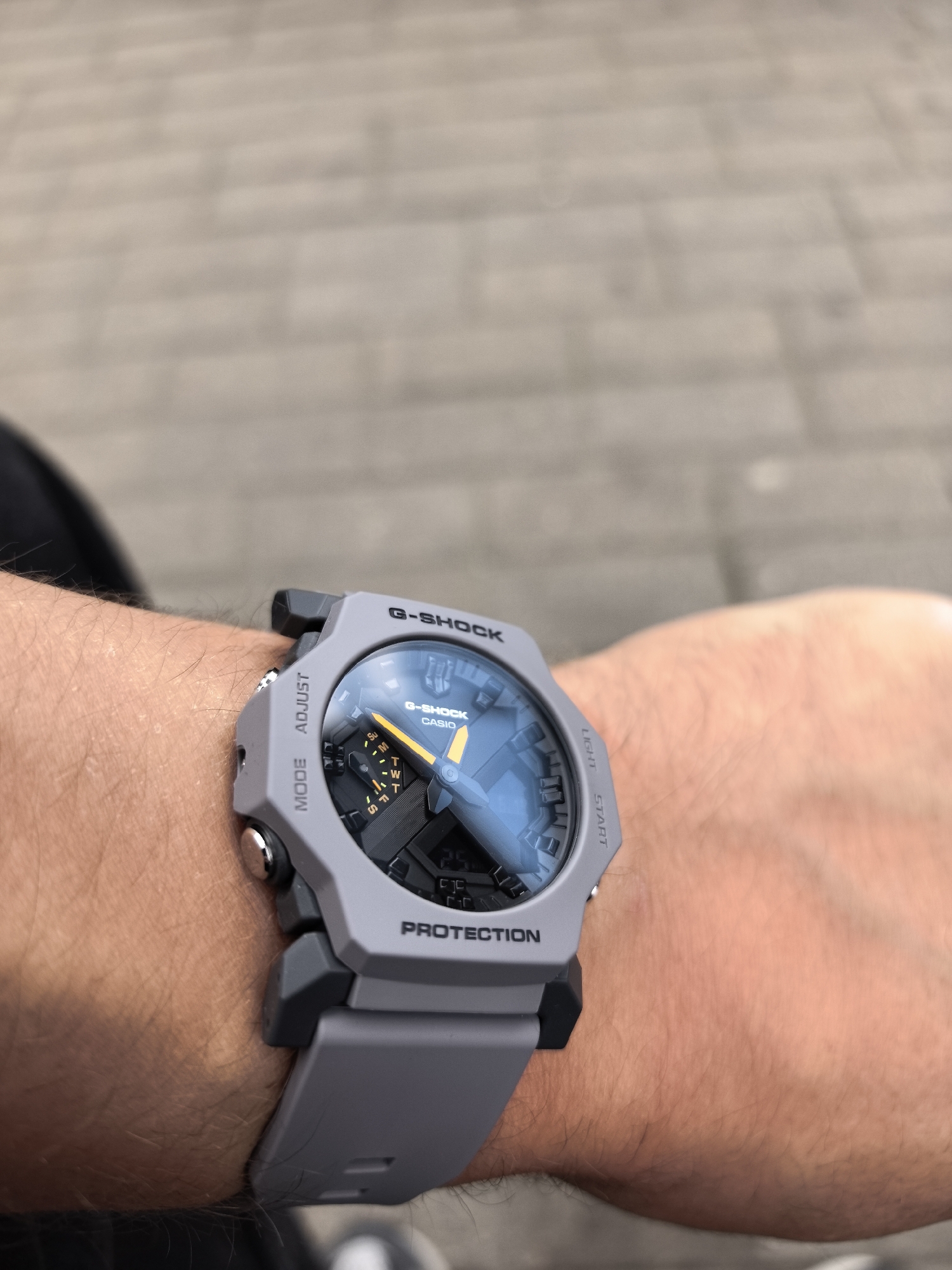 An extreme close-up shot shows a grey G-SHOCK CASIO watch on a person's wrist. The watch has a light blue face with orange accents. The person is wearing a black shirt. The background is out of focus and appears to be a brick-paved walkway. The image was taken in Minsk, Belarus.