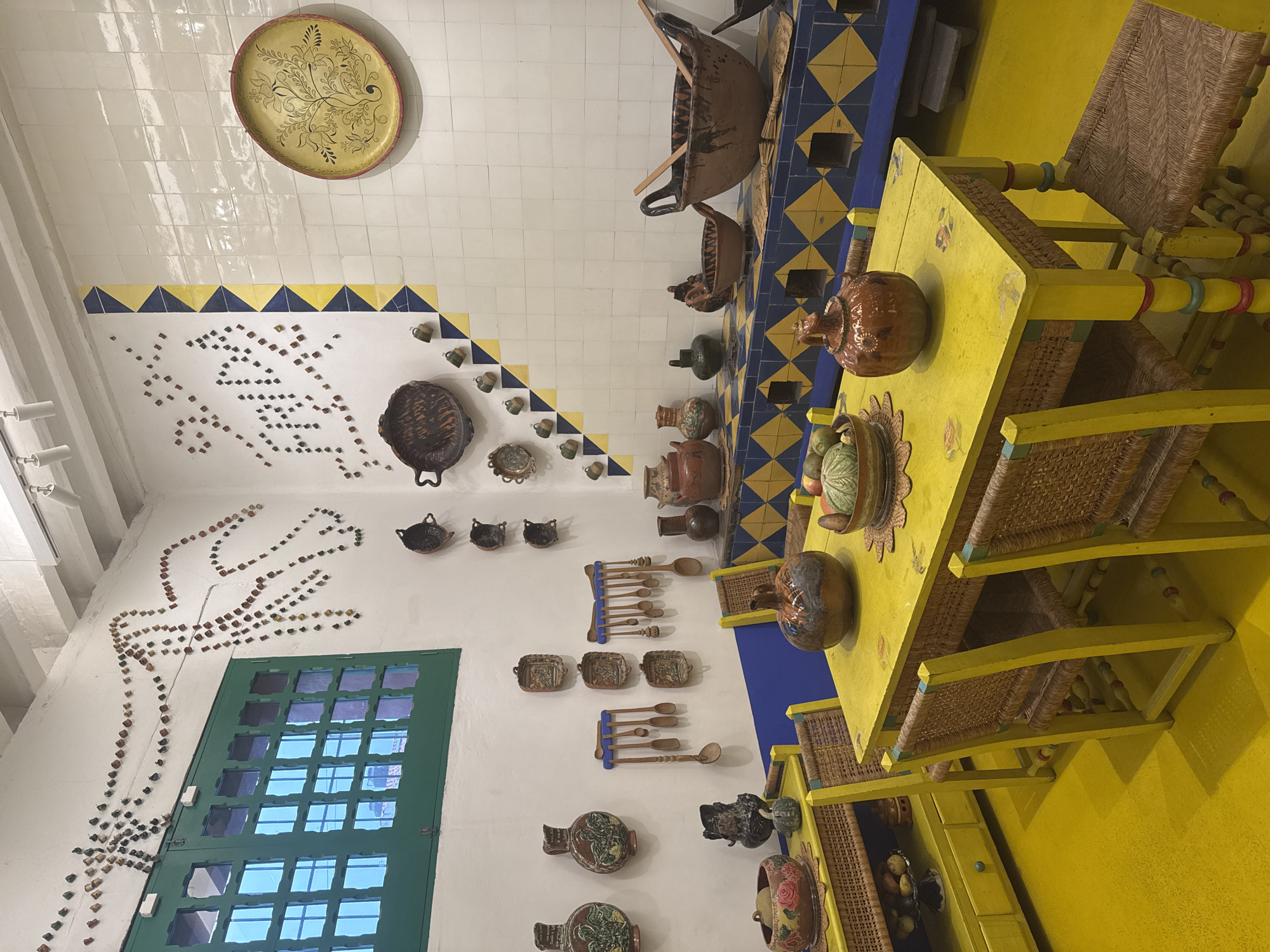 The image captures a colorful, culturally rich interior in Mexico City, Mexico. A table and chairs are prominently featured; the table is painted bright yellow and has a collection of decorative pottery items on top. The chairs, also painted yellow, have woven seats and backs. A striking blue and yellow tiled backsplash adorns a surface behind the table, where a variety of decorative bowls and vessels are displayed. The walls are white, and there is a large green window to the left side of the frame. The wall is adorned with decorative elements, including small objects arranged in the shape of an outstretched hand. A large decorative plate hangs on the wall near the ceiling.