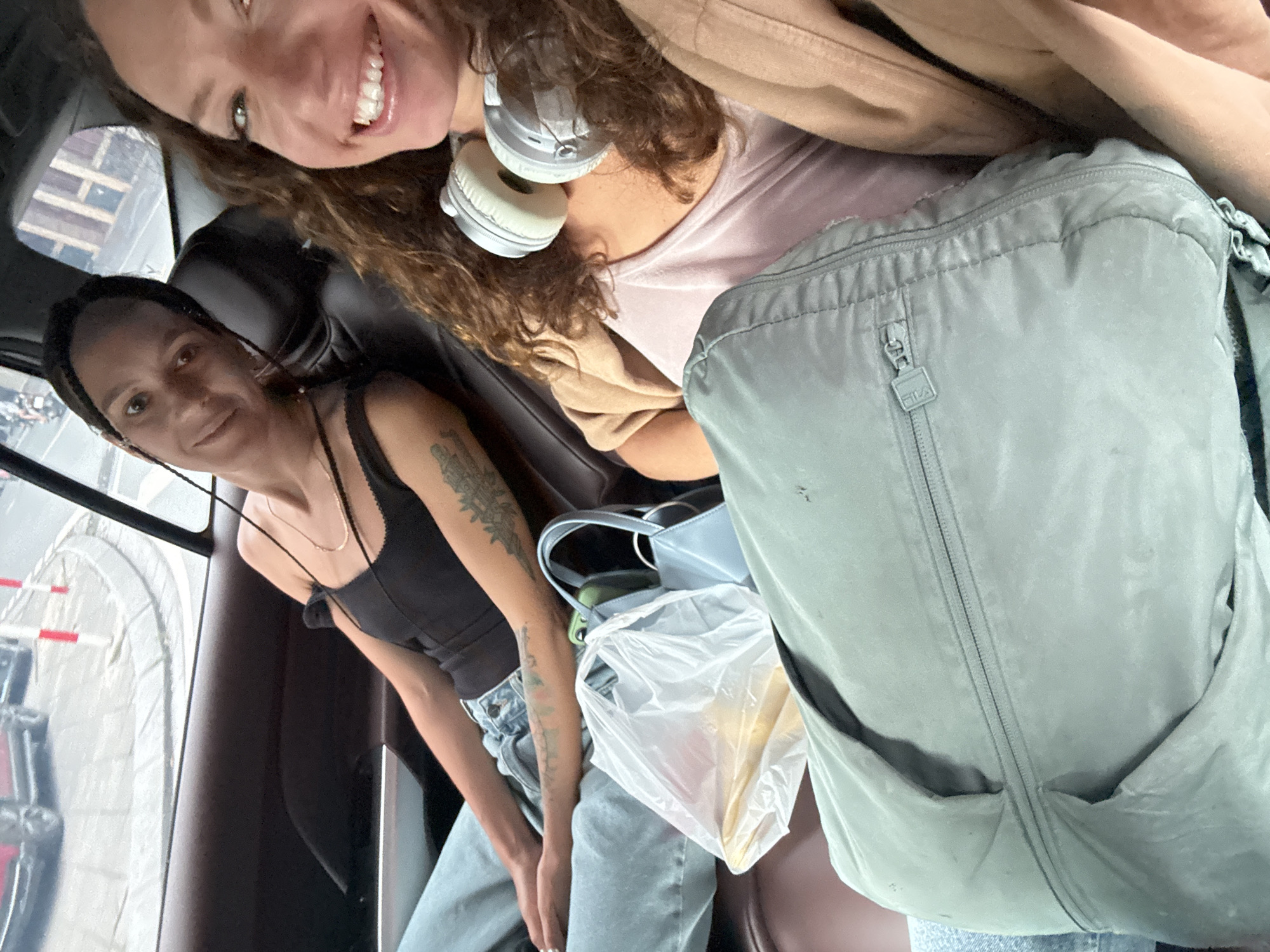 Two women are inside a vehicle, possibly a car, likely a taxi or ride-share, looking at the camera. The woman on the right is smiling broadly with headphones around her neck, wearing a light pink top and a tan jacket. The woman on the left is looking at the camera with a slight smile, wearing a black tank top, jeans, and has tattoos on her arm. Between them is a light blue purse, a clear plastic bag with what appears to be food inside, and a large light grey FILA backpack. Outside the window is a view that suggests the scene is located in Hà Nội, Vietnam. Based on the outside view and road markings, it looks as if the vehicle is driving through or near the elevated section of the Hanoi Ring Road.