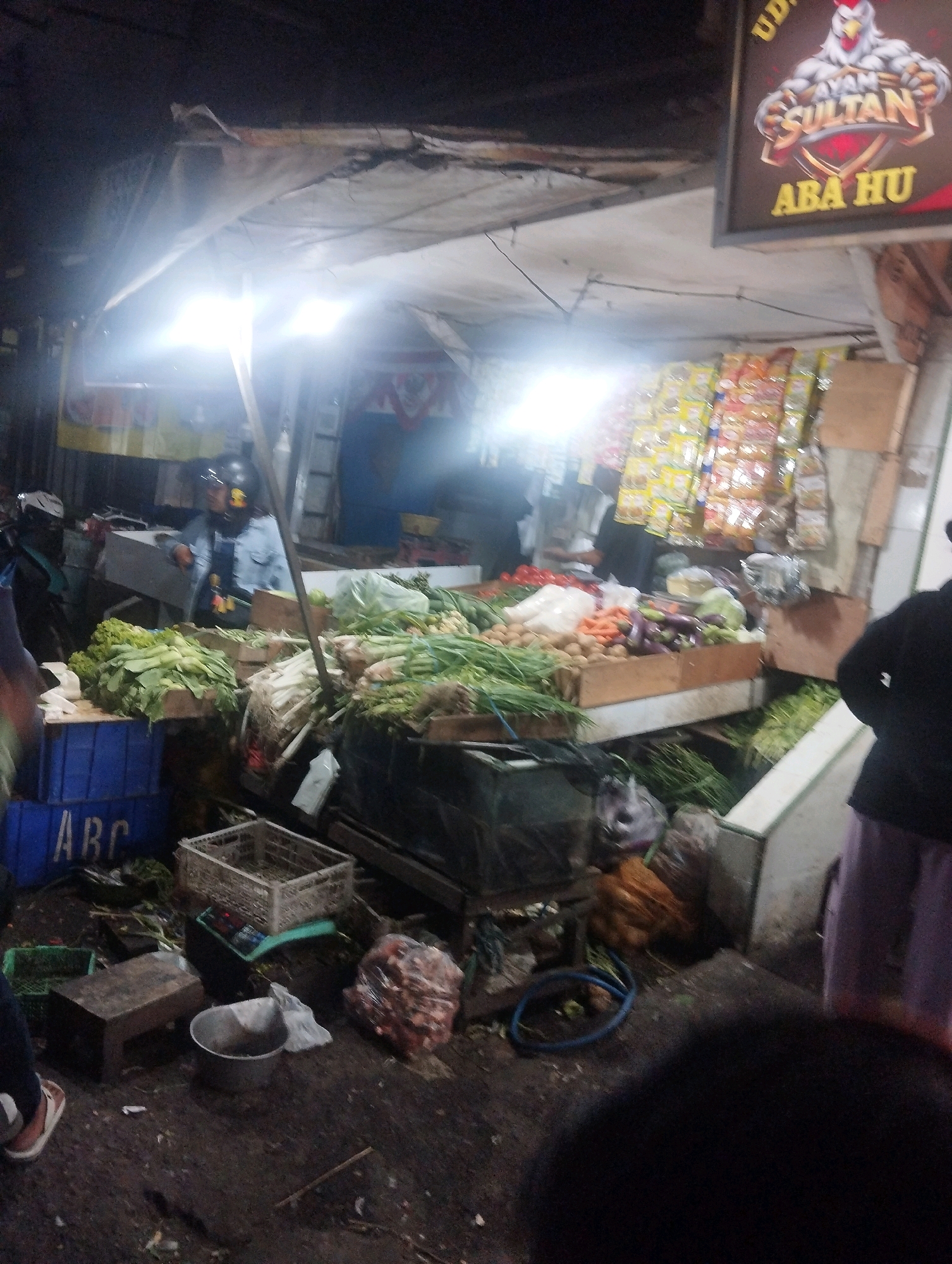 The image shows a nighttime scene at a market stall in the city of Malang, Indonesia. The stall is laden with fresh produce, including leafy greens, onions, tomatoes, carrots, and eggplants. Several blue crates labeled "ABC" are visible, along with other plastic containers and baskets used for displaying and storing goods. A person wearing a helmet and a denim jacket is standing behind the stall, interacting with customers or arranging produce. Another person, partially visible, is wearing a dark jacket and a purple skirt. The market appears to be open-air, with a tarp overhead providing shelter. The lighting is a mix of bright artificial lights from the stall and ambient street lighting, creating a somewhat shadowy atmosphere. The ground is covered in a dark, possibly wet, material. A sign above the stall features a graphic of a rooster and the text "AYAM SULTAN ABA HU", suggesting it is a food stall or a vendor selling poultry-related products. Packaged goods, possibly snacks or seasonings, are hanging on display in the background. The overall impression is of a busy, local market operating after dark.
