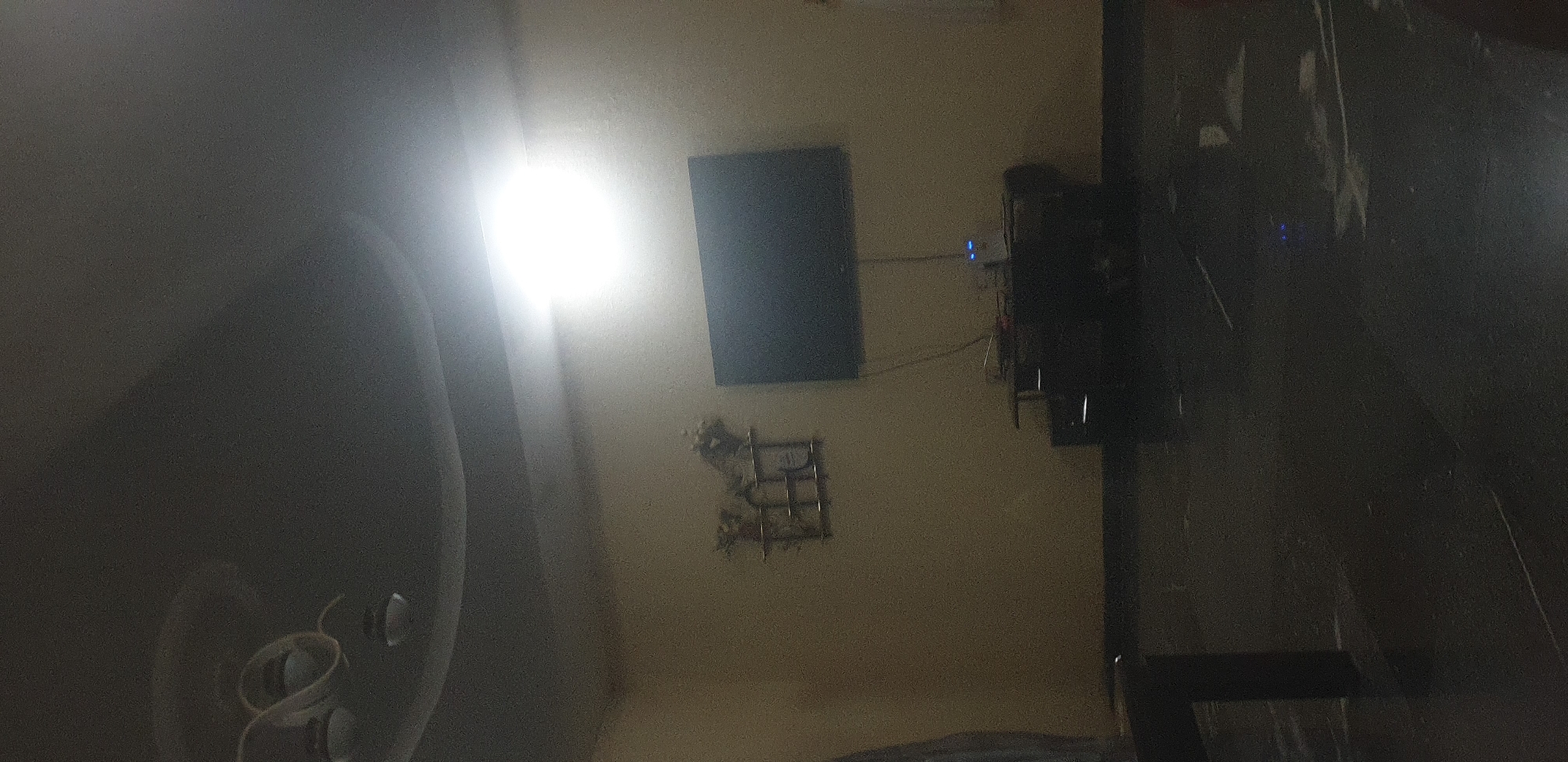 The image shows an indoor scene, likely a living room or entertainment area, in the city of Unknown, Unknown. A flat-screen television is mounted on a wall. Below the television, a dark-colored entertainment stand holds various electronic devices, including what appears to be a router or modem with blue indicator lights. On the wall to the left of the television, a decorative wooden shelf with items placed on it is visible. The ceiling features recessed lighting and a light fixture. The floor is dark and appears to be tiled or polished. The overall lighting in the room is dim, with a bright light source coming from the ceiling, creating a spotlight effect on the wall and partially illuminating the scene. There are no people visible in the image.