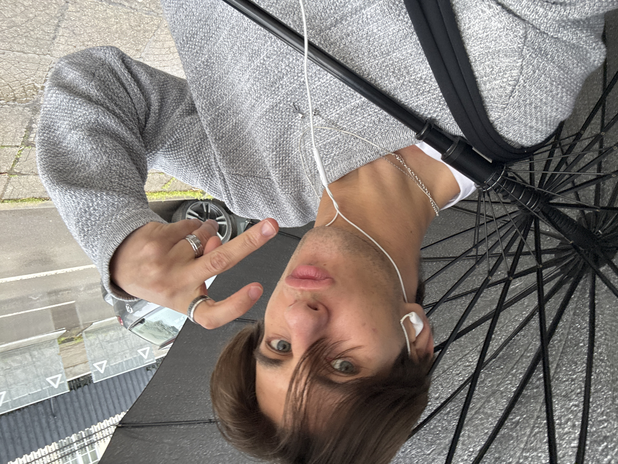 A young man takes a selfie outdoors in Sofia, Bulgaria, captured from a top-down perspective that makes the image appear rotated. He is looking directly at the camera with greenish-blue eyes and pouting lips, holding up two fingers in a peace sign with his left hand. His dark brown hair is styled with bangs falling across his forehead.
He is wearing a grey, textured sweater over a white t-shirt, and accessorized with a silver chain necklace and multiple rings on his left hand (two on the index finger, one on the ring finger). White wired earbuds are visible, suggesting he is listening to audio.
He is holding a black umbrella, which is partially open and seen from underneath, with its dark spokes visible. This, combined with the damp-looking paved ground, suggests overcast or rainy weather. The background features a paved surface, possibly a sidewalk or street, with small patches of greenery. A grey car is partially visible behind him. Further back, white triangular road markings can be seen on an asphalt surface, possibly indicating a pedestrian crossing or specific road rules. There is no visible text in the image.