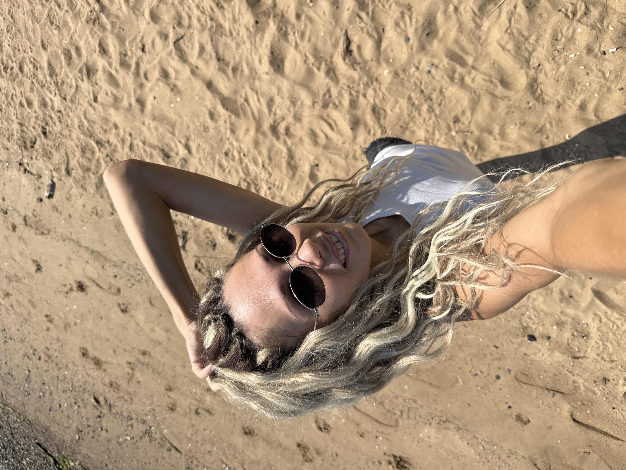 The media file shows a person lying on a sandy surface, likely a beach or dune, with a top-down perspective. The individual has blonde, wavy hair and is wearing sunglasses and a white top. Their face is partially visible, smiling, and they appear relaxed. One arm is raised above their head, with the hand resting on their hair, while the other arm is also extended, possibly holding the camera. The sandy ground is textured with footprints and small debris like shells and pebbles. Sunlight casts a shadow from the person onto the sand, suggesting it's a sunny day. The overall scene conveys a sense of leisure and enjoyment. The provided location context is Rusanovo, Russia.