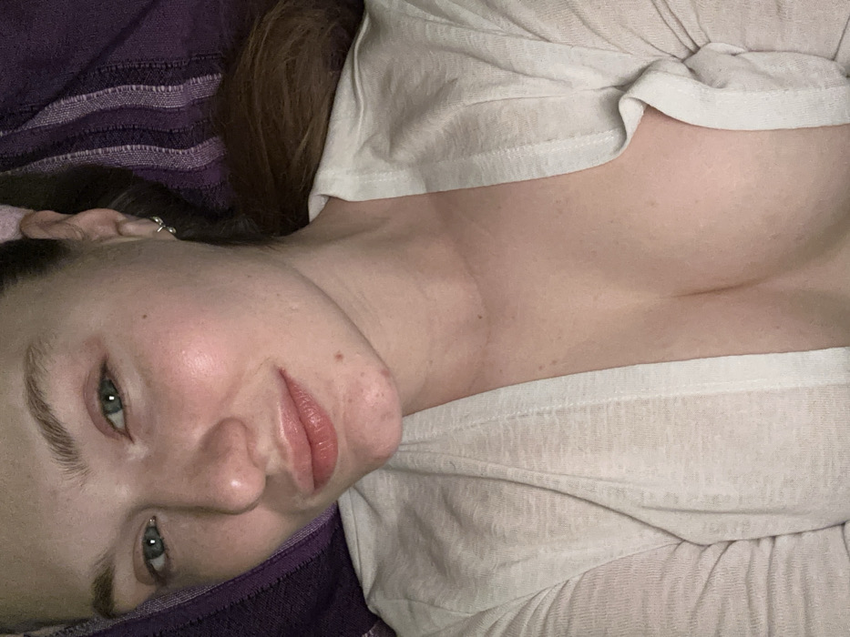 A woman reclines on a purple striped surface, her head tilted towards the camera. Her gaze is direct, revealing light-colored eyes and pink lips. She wears a loose-fitting, light-colored top that exposes part of her chest. A small earring is visible in her left ear. Her hair is a dark shade, contrasting with her fair skin. The overall lighting is soft, highlighting her features and creating a relaxed atmosphere.