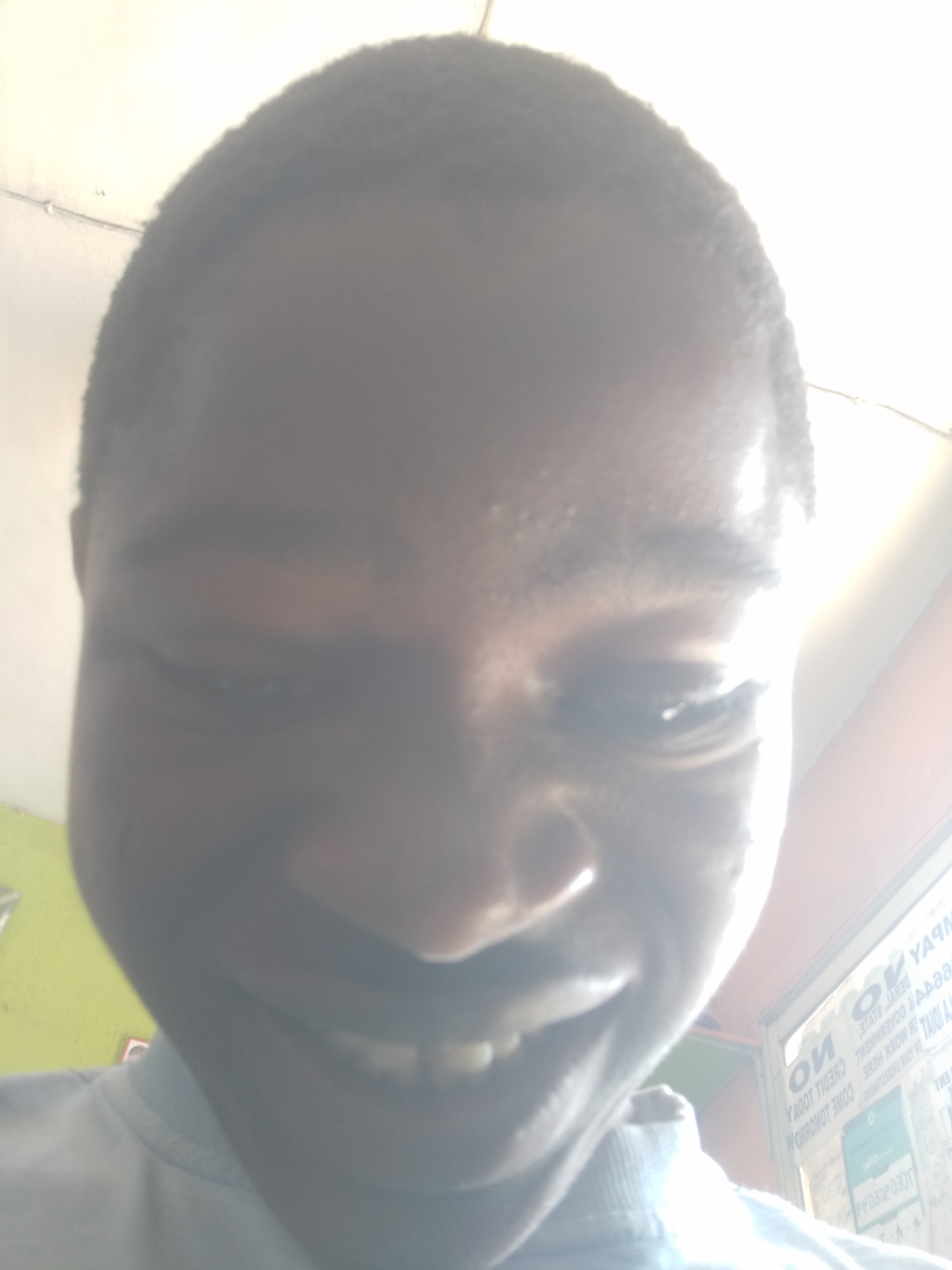 A close-up, low-angle selfie of a Black man with a shaved head, smiling slightly, with a bright light source above him creating significant glare. The background is partially obscured but shows a lime green wall to the left and a partially visible, text-covered surface, possibly a window or wall, to the right. The text appears reversed, and the lighting suggests the photo was taken indoors. Without more visual clues, it's impossible to confirm the location as Shangisha, Nigeria, or any specific detail regarding its environment.